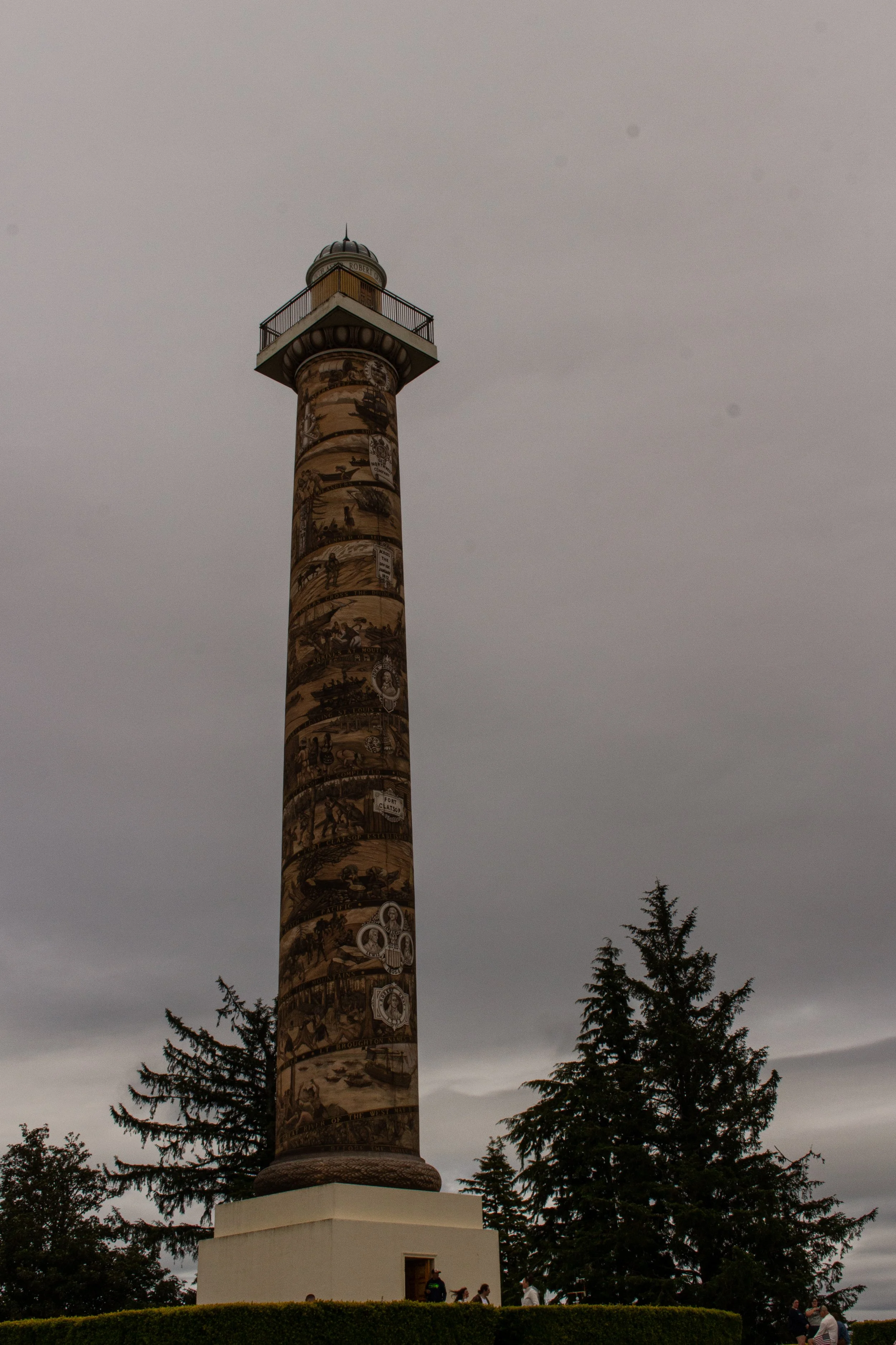 A tall monument with historical murals or illustrations on its surface, topped with a small structure, and surrounded by trees and a cloudy sky.