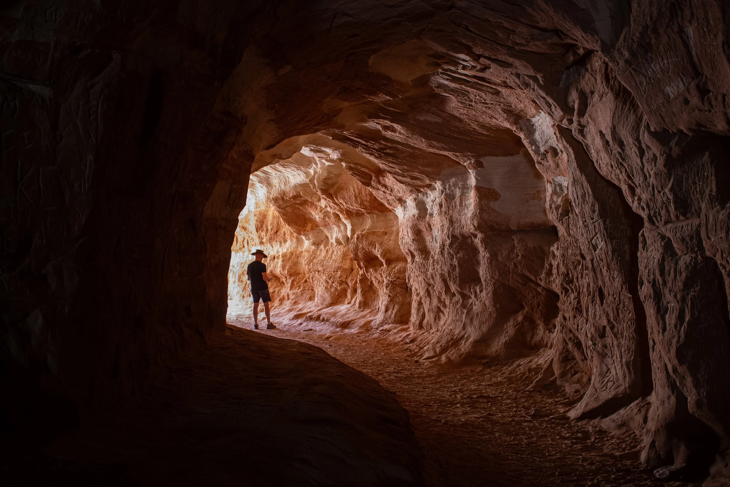 Person standing inside a large, reddish-brown cave, illuminated by natural light coming from the outside or another opening, with textured rock walls surrounding them.