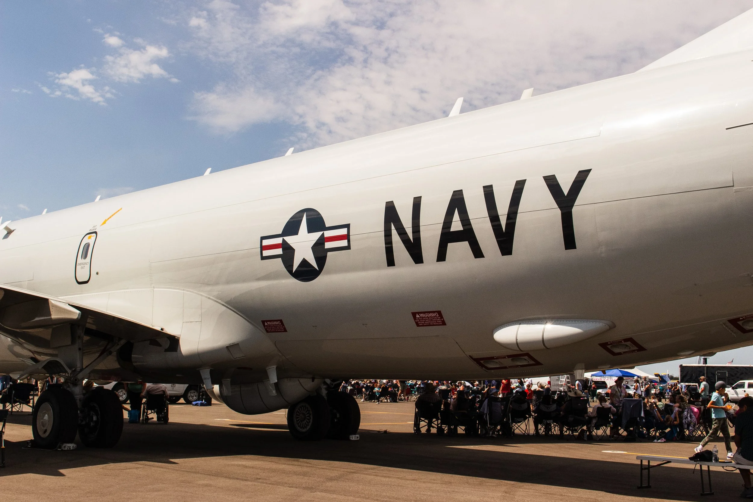 A large aircraft with the words 'NAVY' and a star insignia on its side is displayed at an outdoor event, with crowds of people sitting and walking around underneath and nearby, and a partly cloudy sky above.