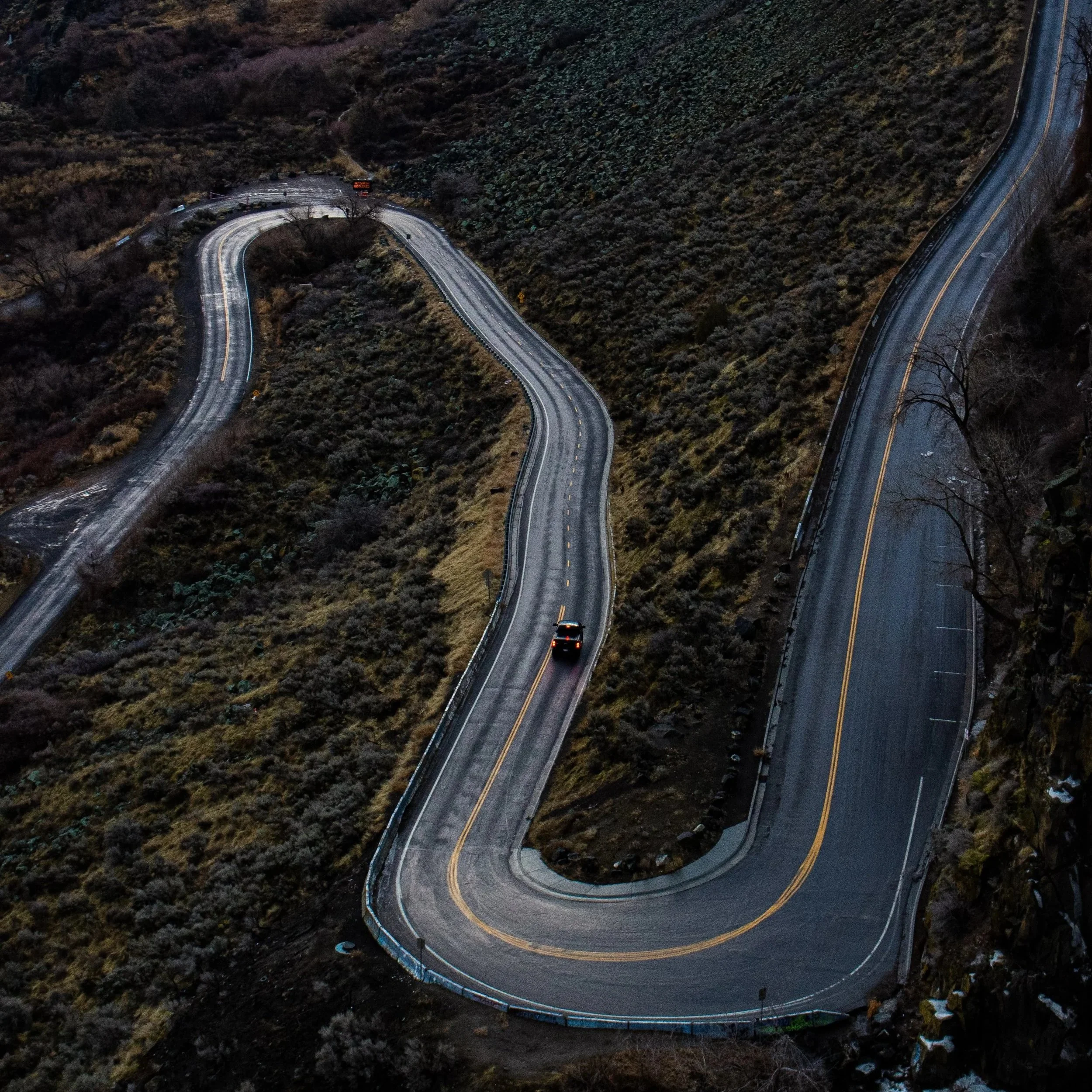 An aerial view of a winding mountain road at dusk, with two cars traveling and displaying streaks of light from their headlights.