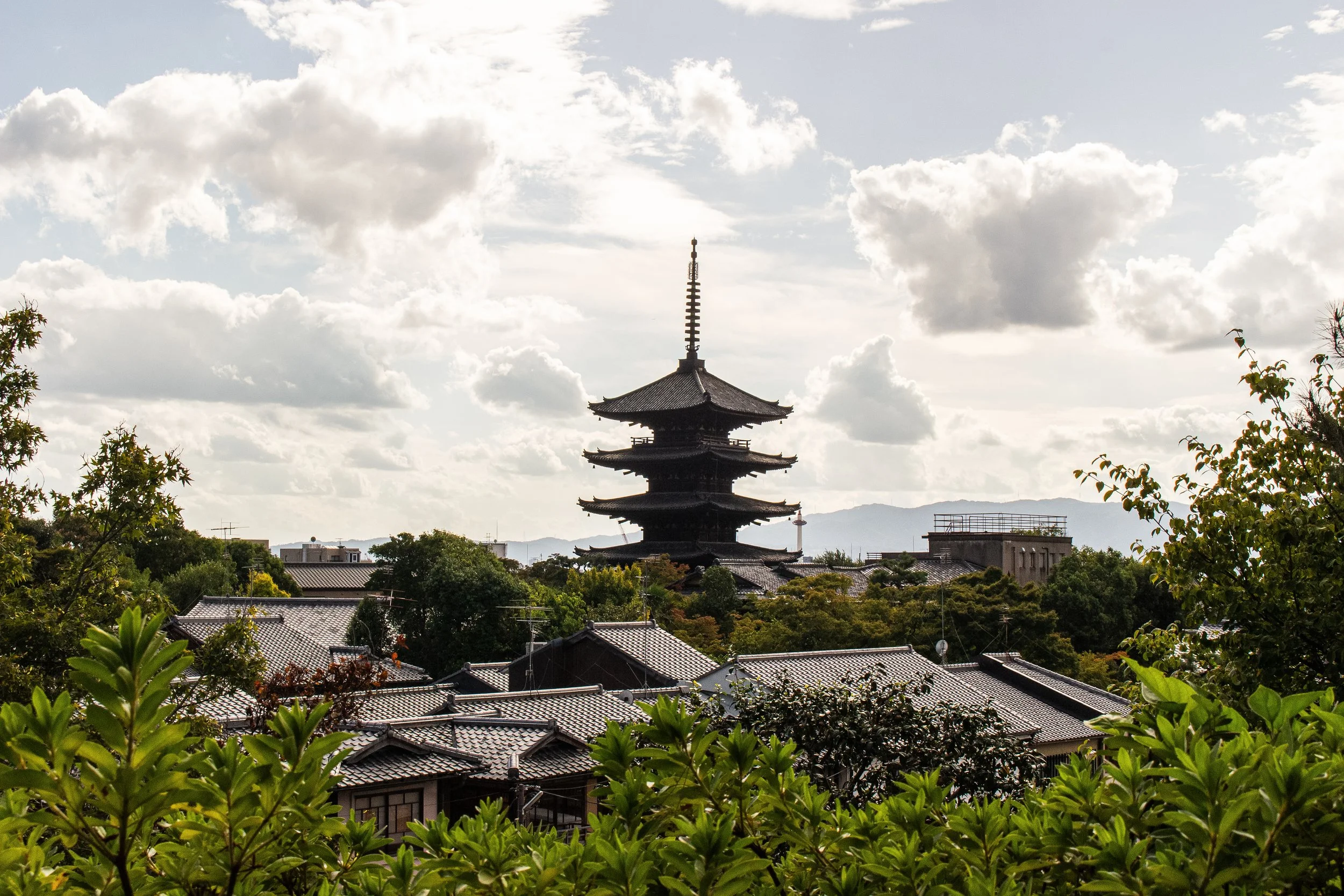 Traditional Japanese pagoda surrounded by greenery with rooftops in the foreground and a cloudy sky in the background.