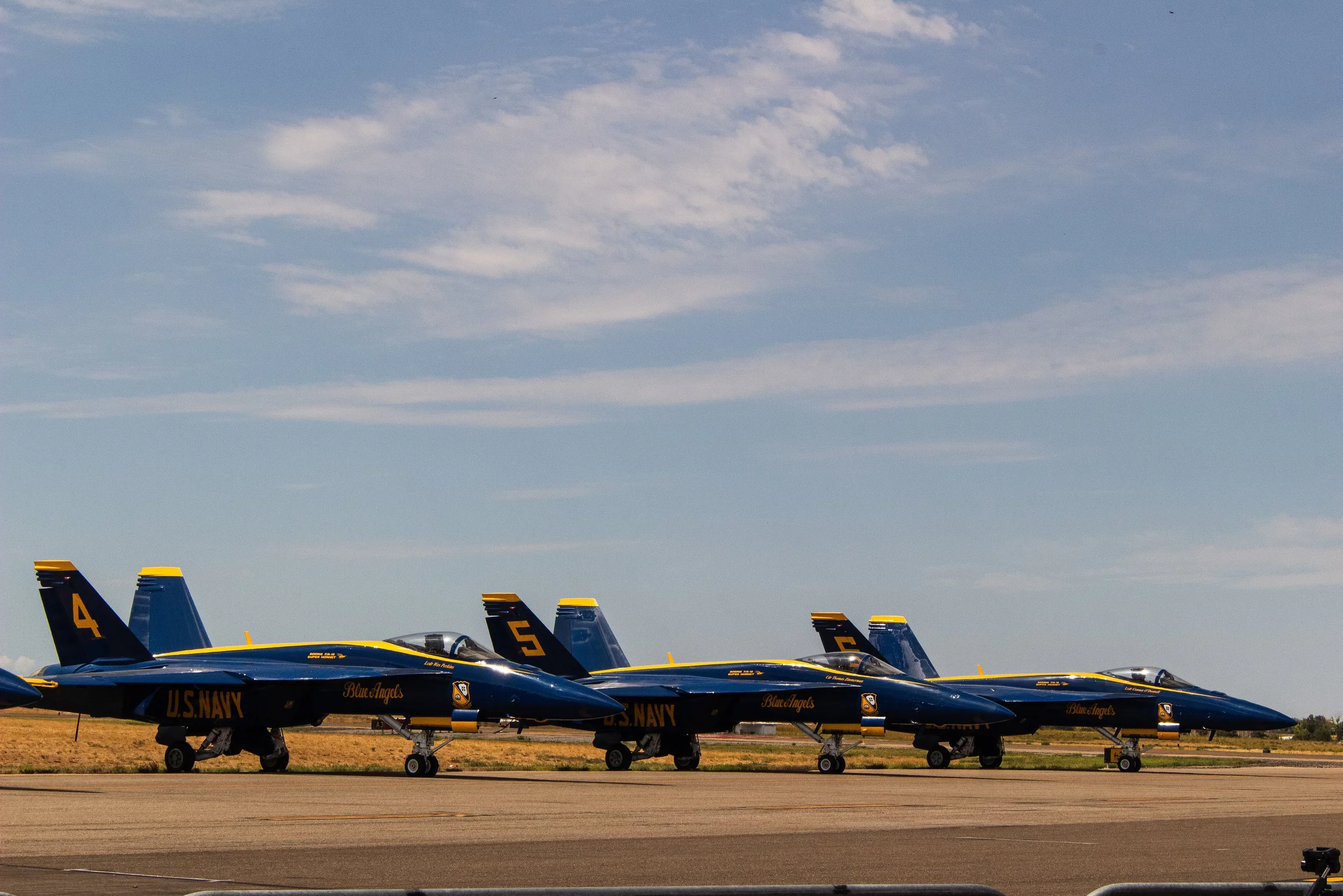 Several Blue Angels military jet fighter planes with navy blue and yellow markings are lined up on the runway, with a clear blue sky and some clouds above.