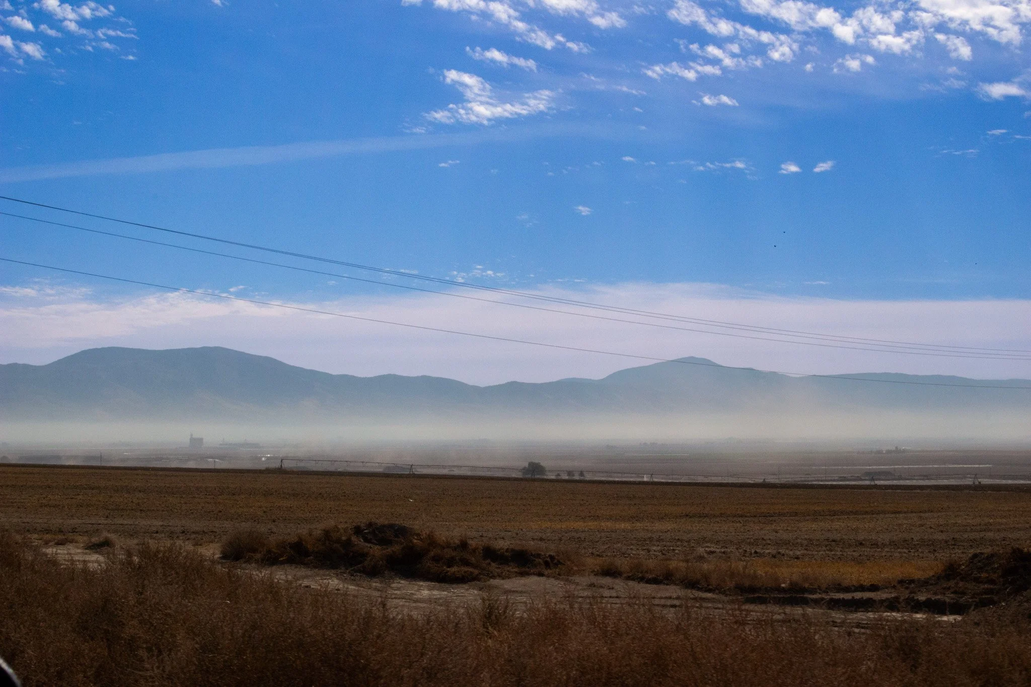 A landscape with mountains in the background, a layer of fog, and flat farmland in the foreground. Power lines cross the sky, which is partly cloudy with scattered clouds.