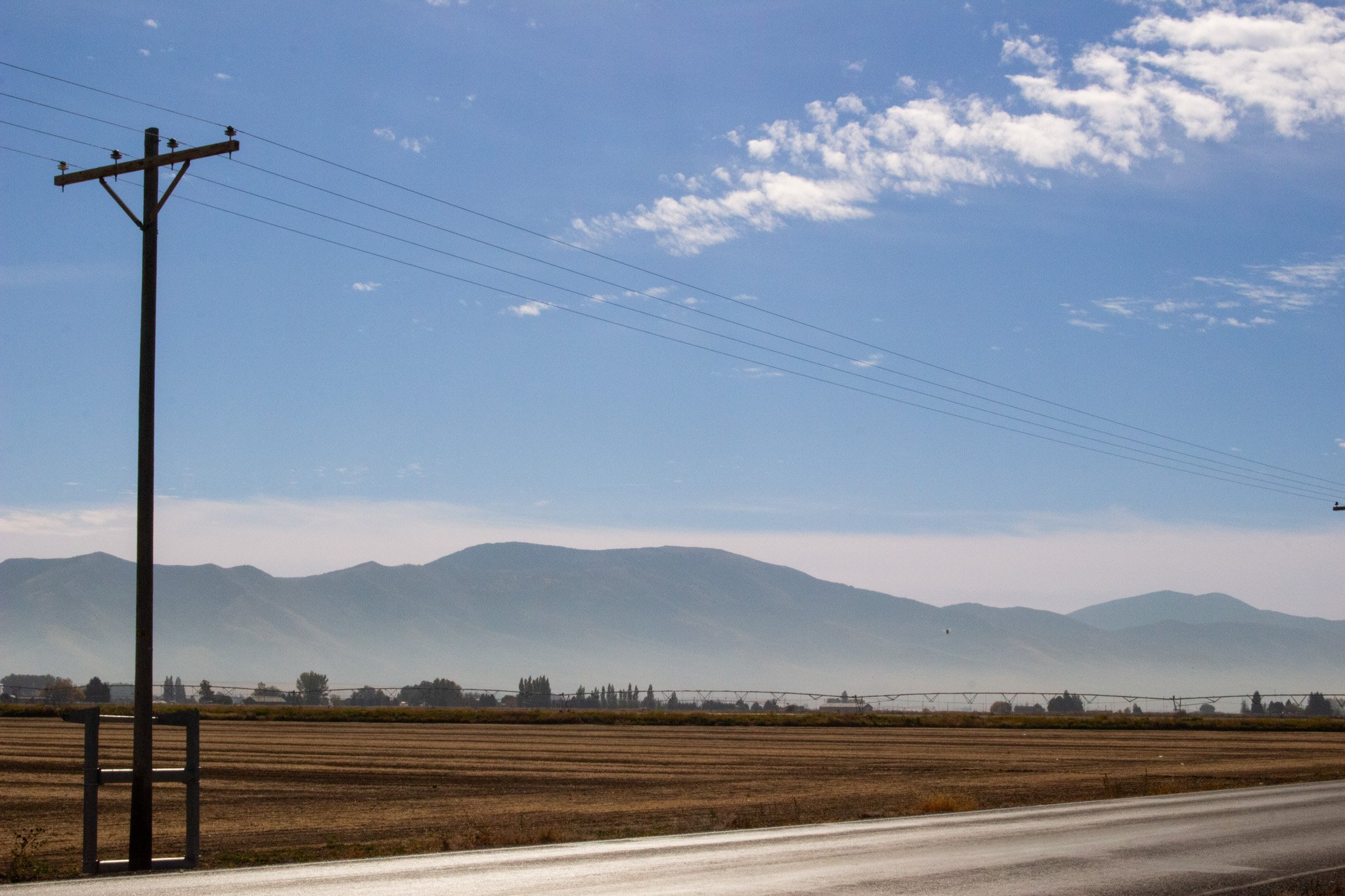 A landscape scene with a utility pole and power lines in the foreground, a plowed field, distant mountains, and a partly cloudy blue sky.