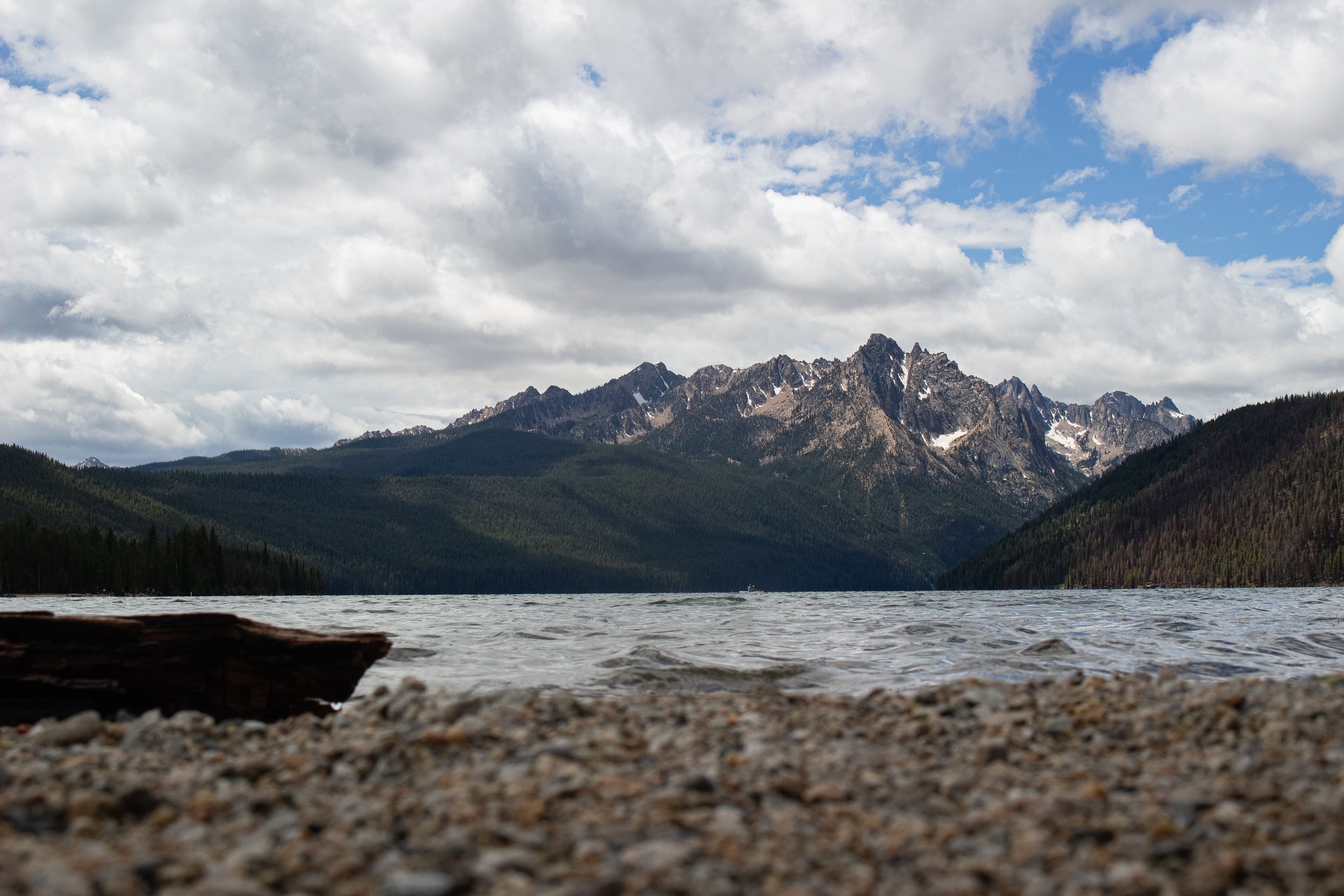 A scenic landscape of a mountain range with snow-capped peaks, a forested valley, and a lake in the foreground under a partly cloudy sky.