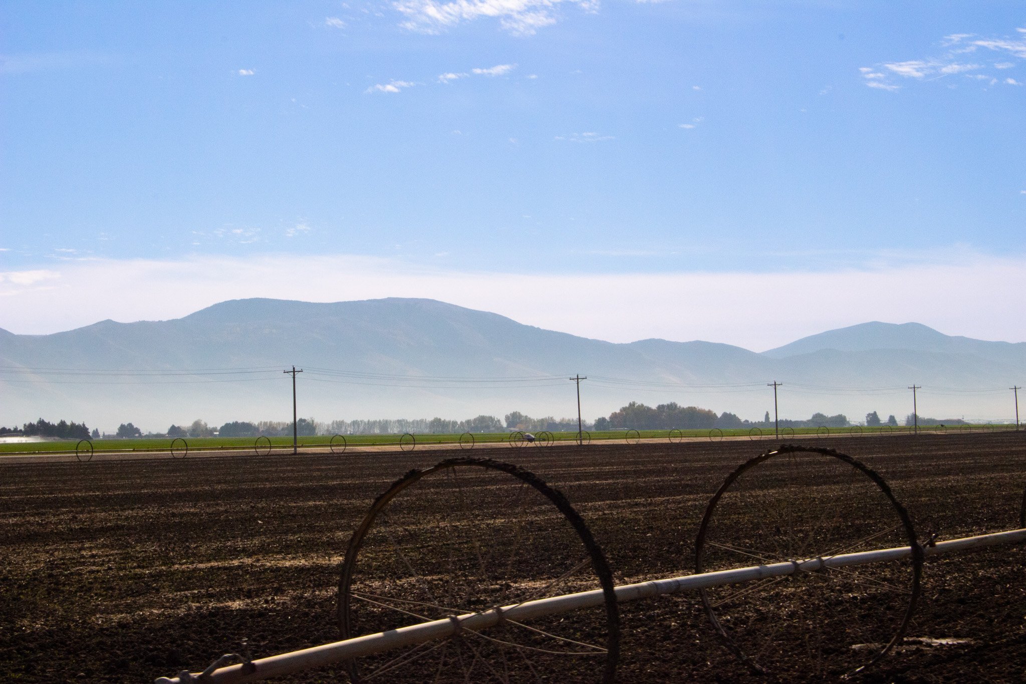 Wide view of a rural landscape with a dark brown plowed field in the foreground, power lines stretching across the middle ground, and a range of mountains in the background under a mostly clear sky with some scattered clouds.