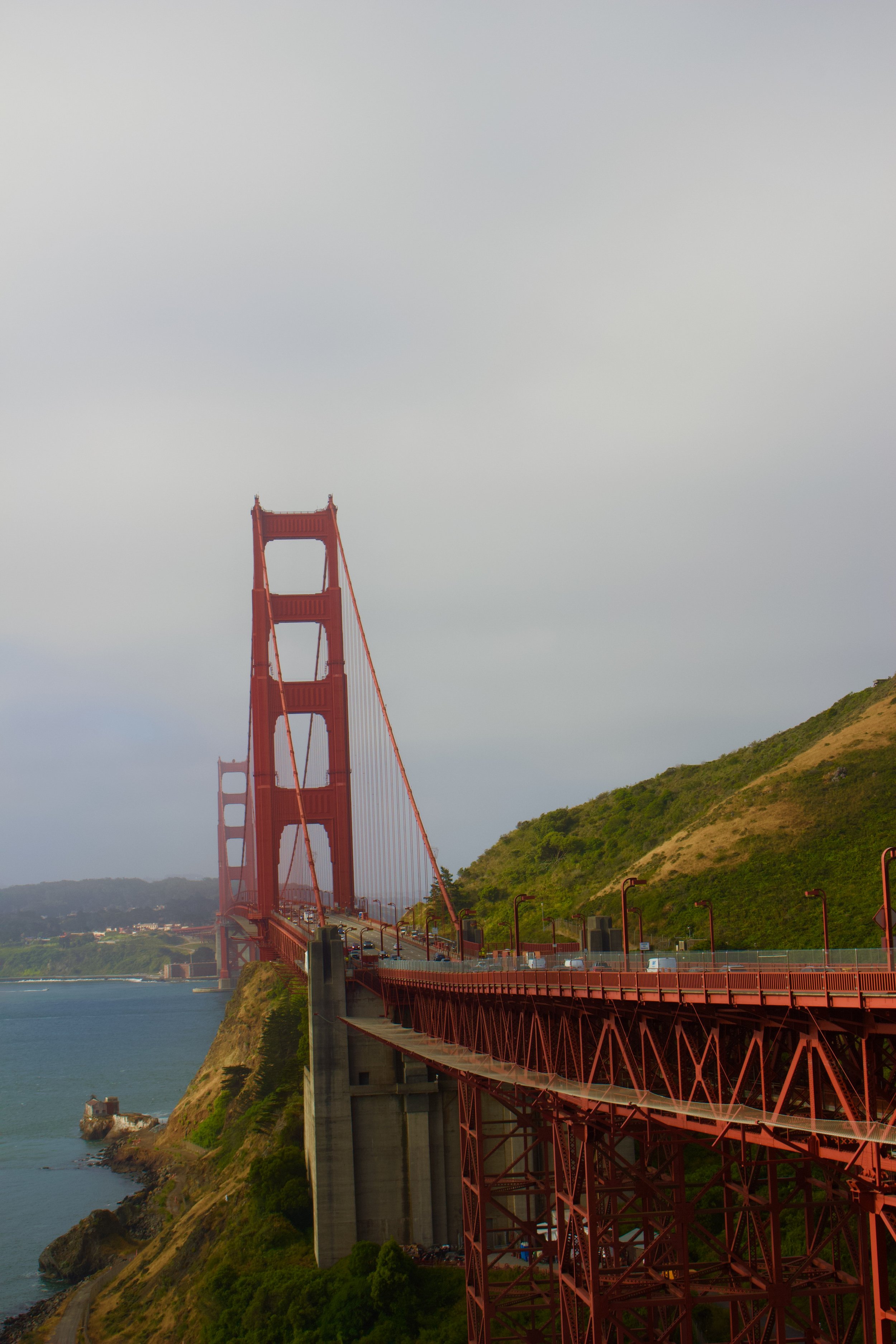 The Golden Gate Bridge in San Francisco, California, seen from a vantage point on the south side with a view of the bridge's red towers, suspension cables, and surrounding hilly landscape with greenery and water below.