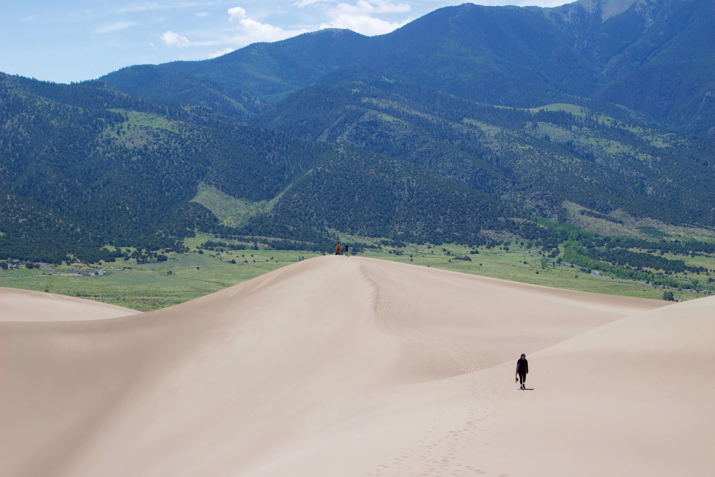Sand dunes in the foreground with two people walking on them, green fields and trees in the middle ground, and large green mountains under a partly cloudy sky in the background.