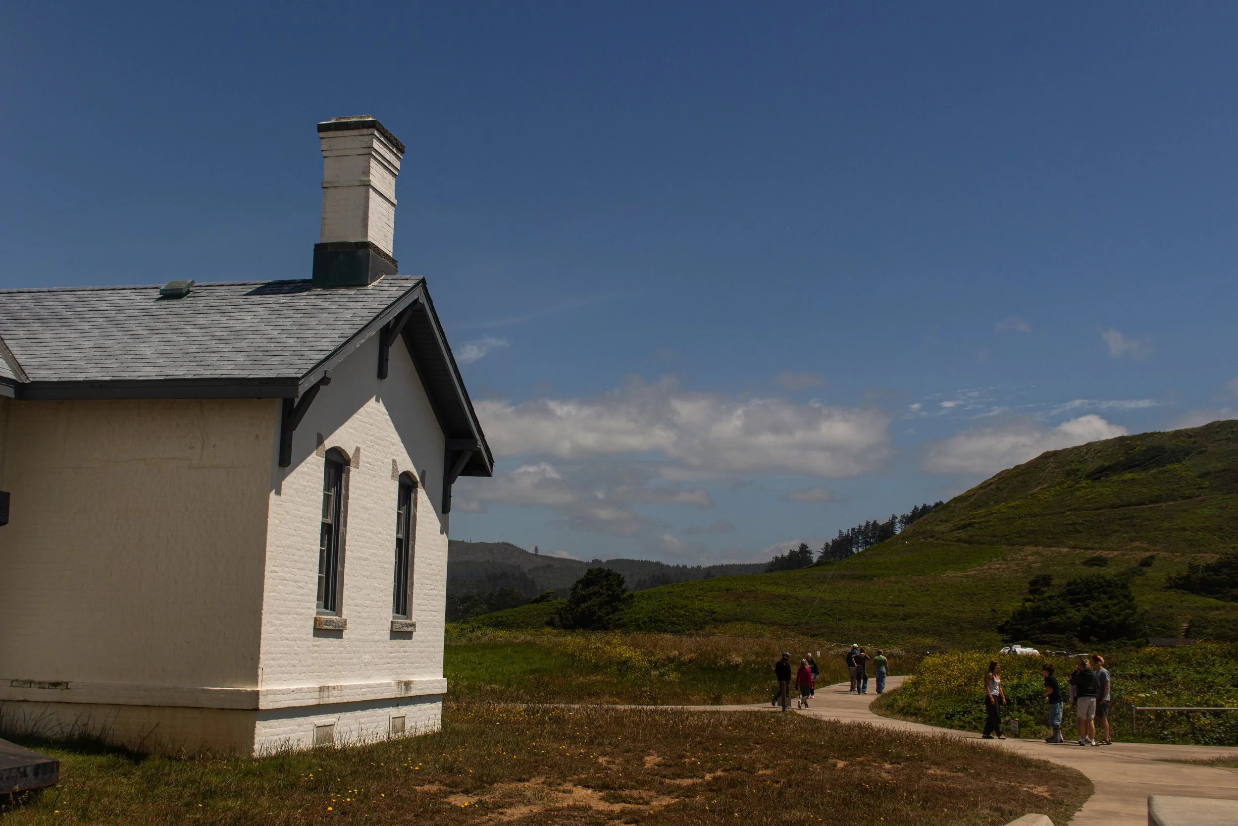 A white house with a chimney on a grassy hill under a blue sky with some clouds. Several people are walking along a paved path near the house.