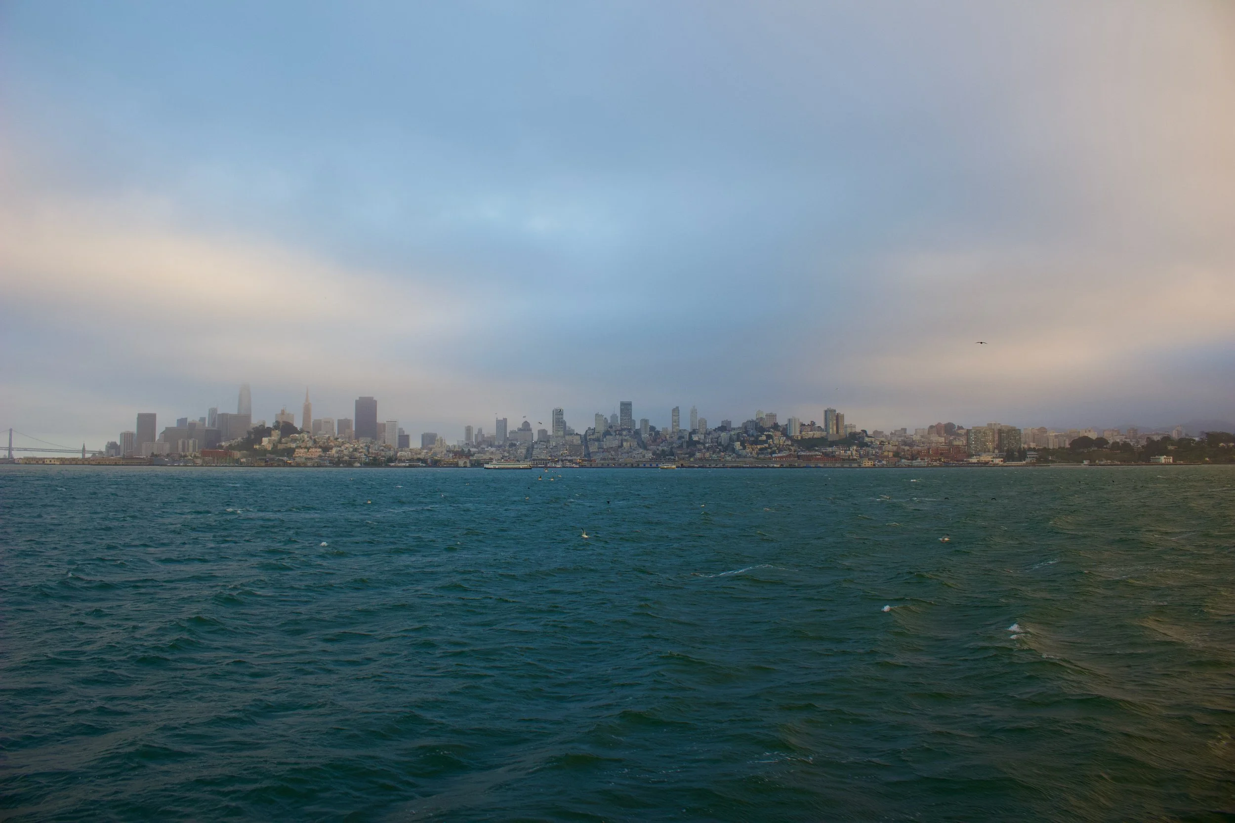A city skyline viewed from across a body of water, with a large bridge on the left and the city buildings shrouded in fog or haze.