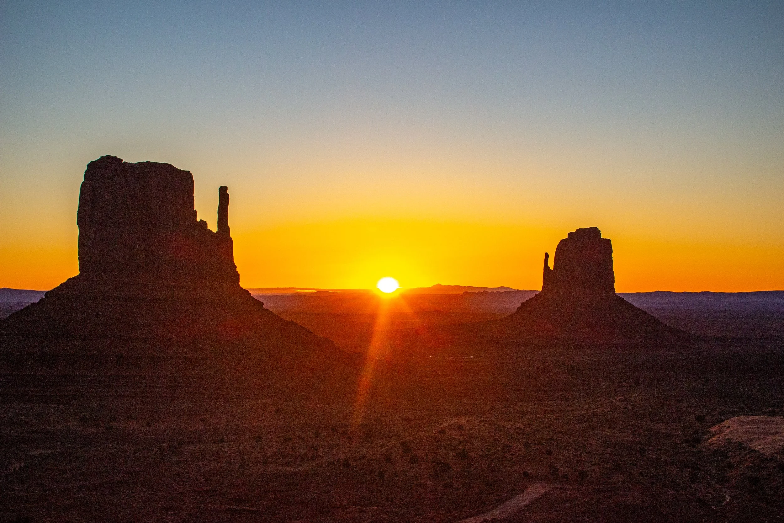 Silhouettes of two large rock formations at sunset with a bright sun near the horizon and a clear sky.