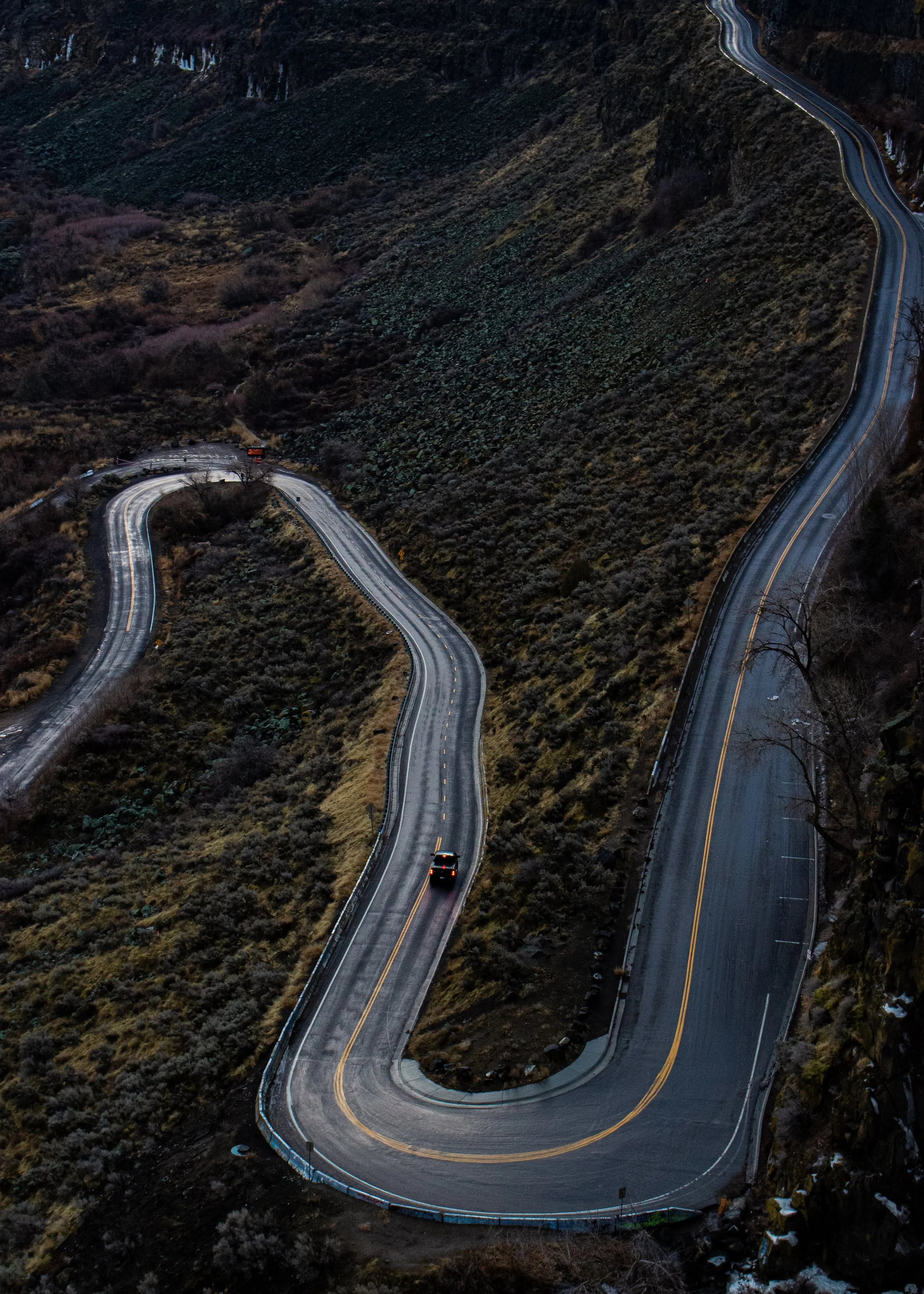 Photo of a mountain road with a sharp switchback, showing a black vehicle driving through the curve surrounded by hilly terrain and sparse vegetation.