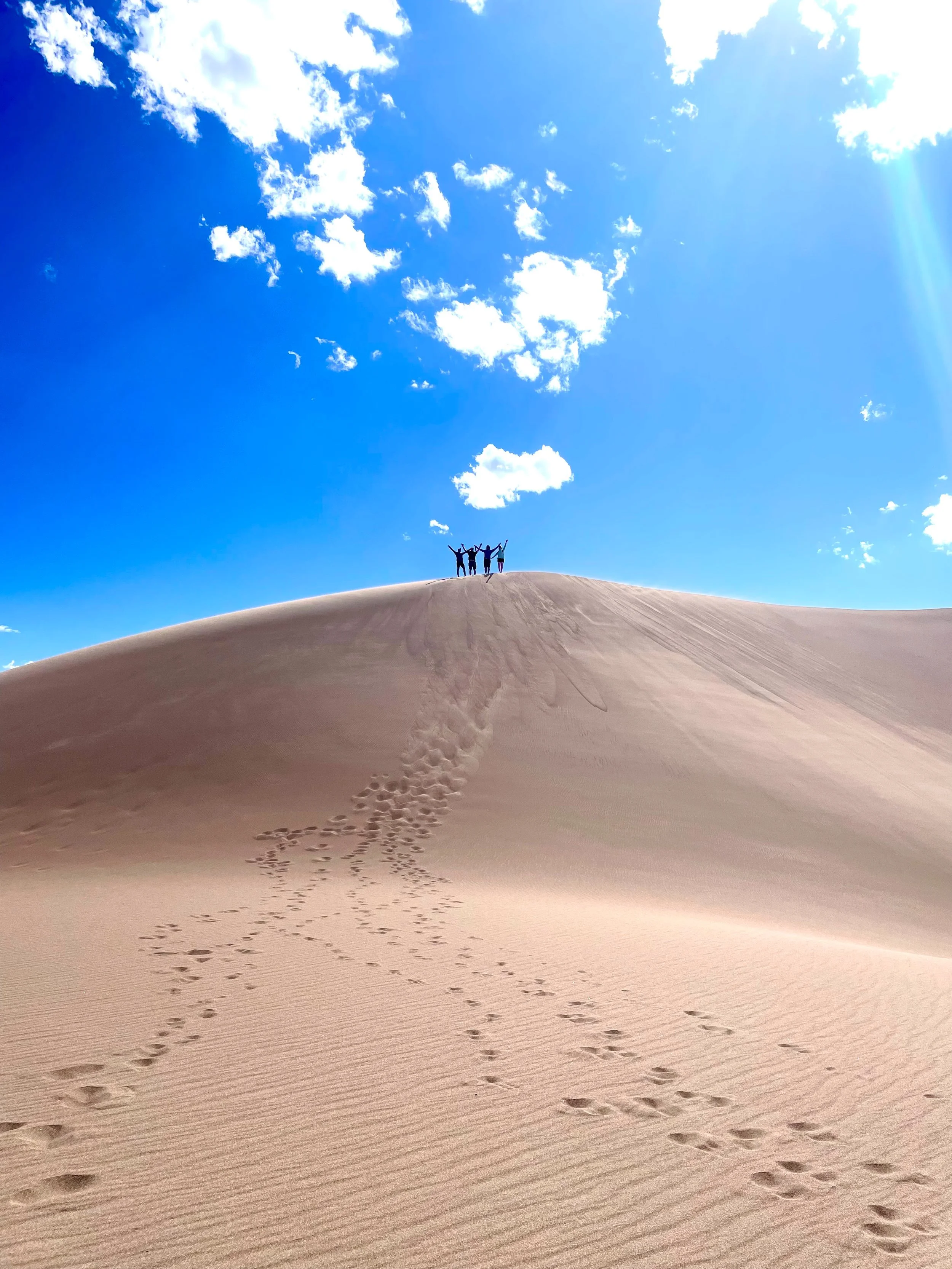 Footprints in the sand leading up a sand dune, with four people at the top with arms raised in the desert under a blue sky with scattered clouds.