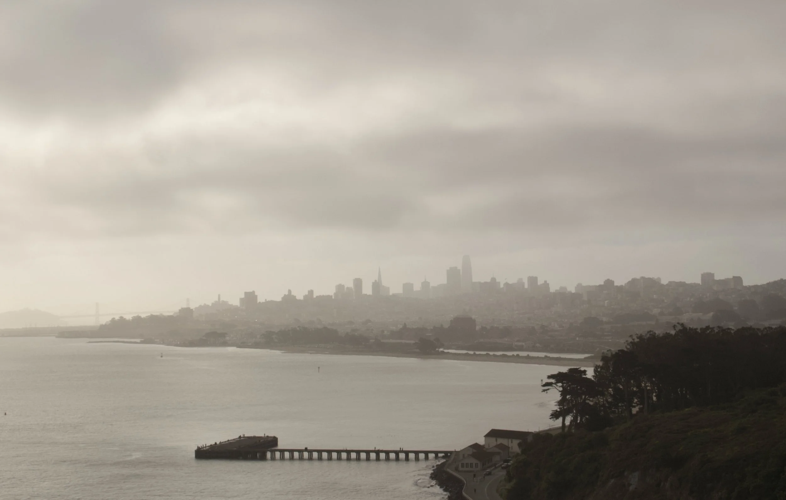 Gray cloudy sky over a city skyline with a body of water in the foreground, including a pier extending into the water and some houses along the shoreline.