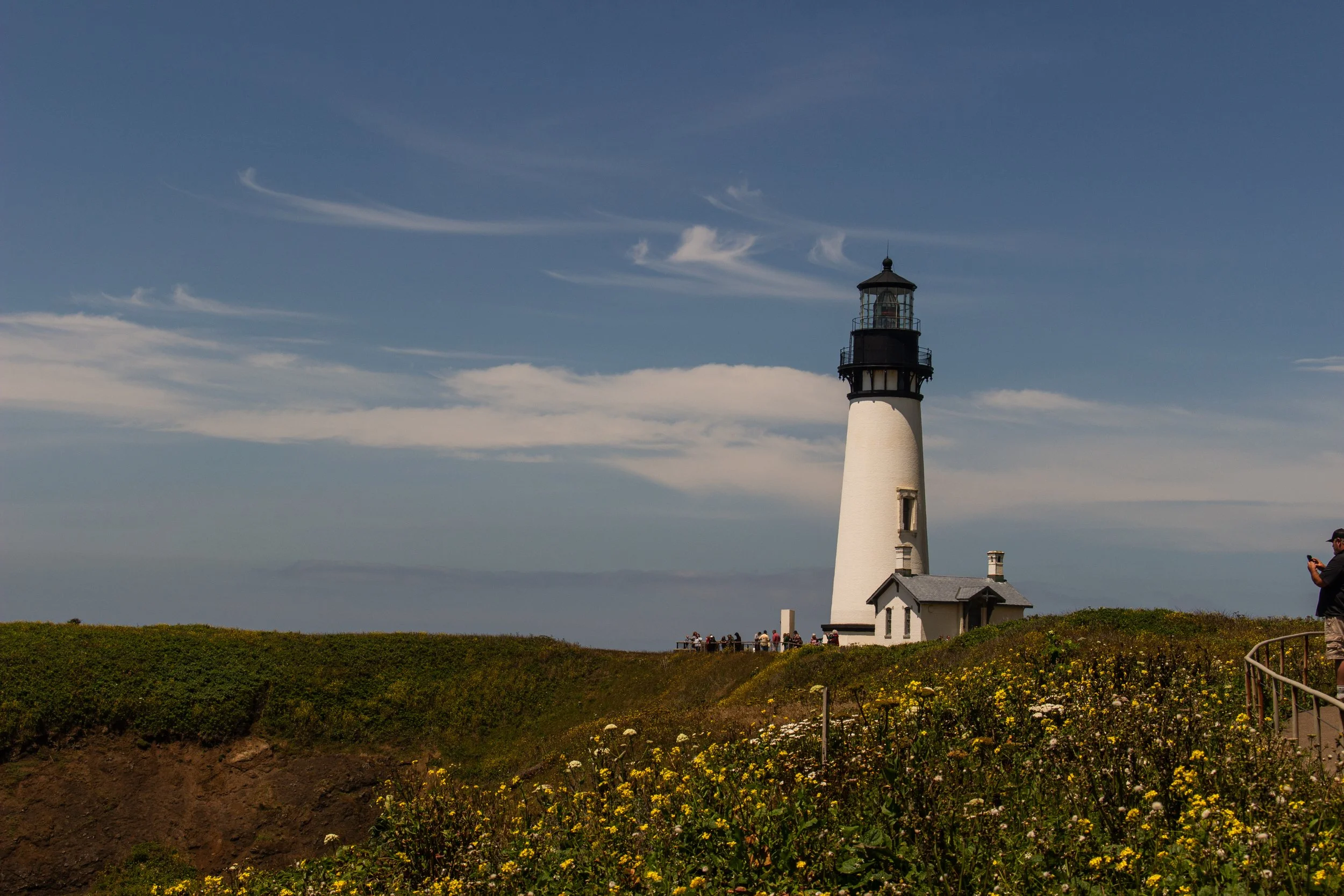 A white lighthouse with a black top on a grassy area with flowers, under a blue sky with wispy clouds. People are near the lighthouse, and someone is taking a photo.