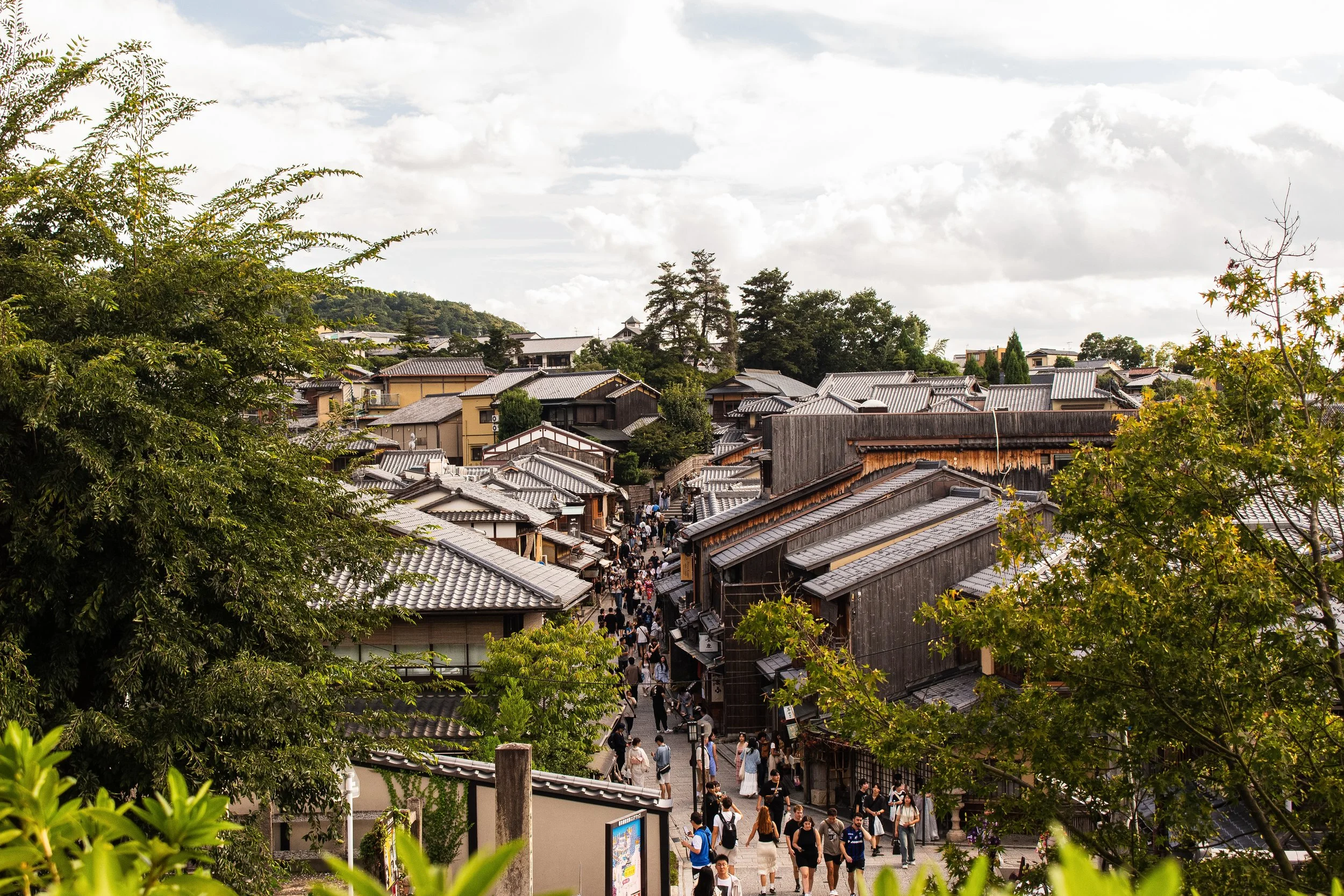 A busy street in a traditional Japanese town lined with wooden buildings and shops, with people walking and browsing. The scene is framed by green trees and the sky is partly cloudy.
