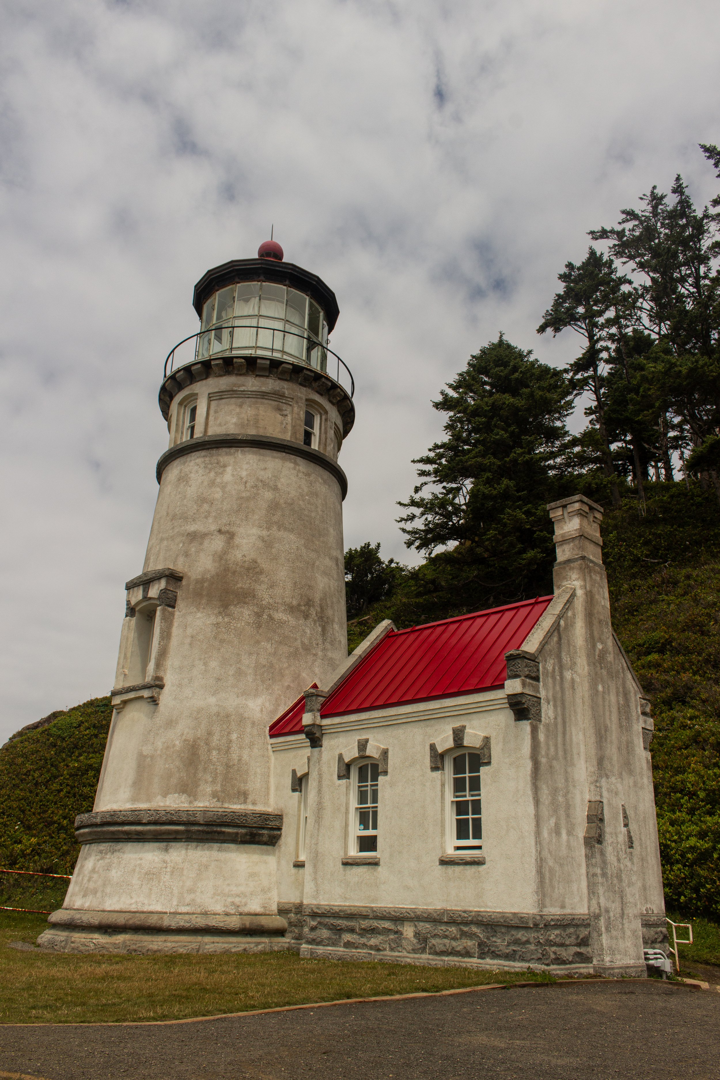 A historic lighthouse with a cylindrical tower and a red roofed building attached, situated in a lush green area near a cloudy sky.