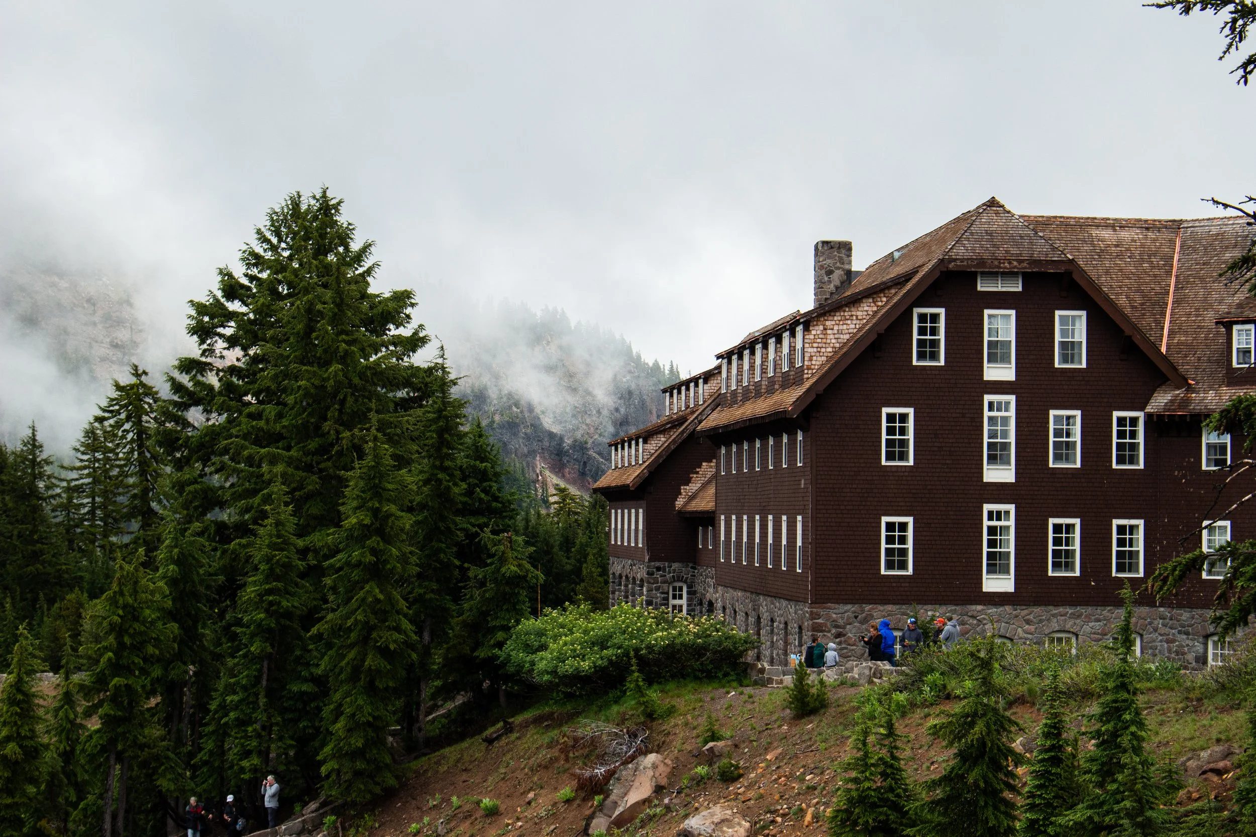 A mountain lodge with brown siding and numerous windows surrounded by lush green pine trees, with foggy mountain hills in the background, and a group of people gathered near the building's stone foundation.