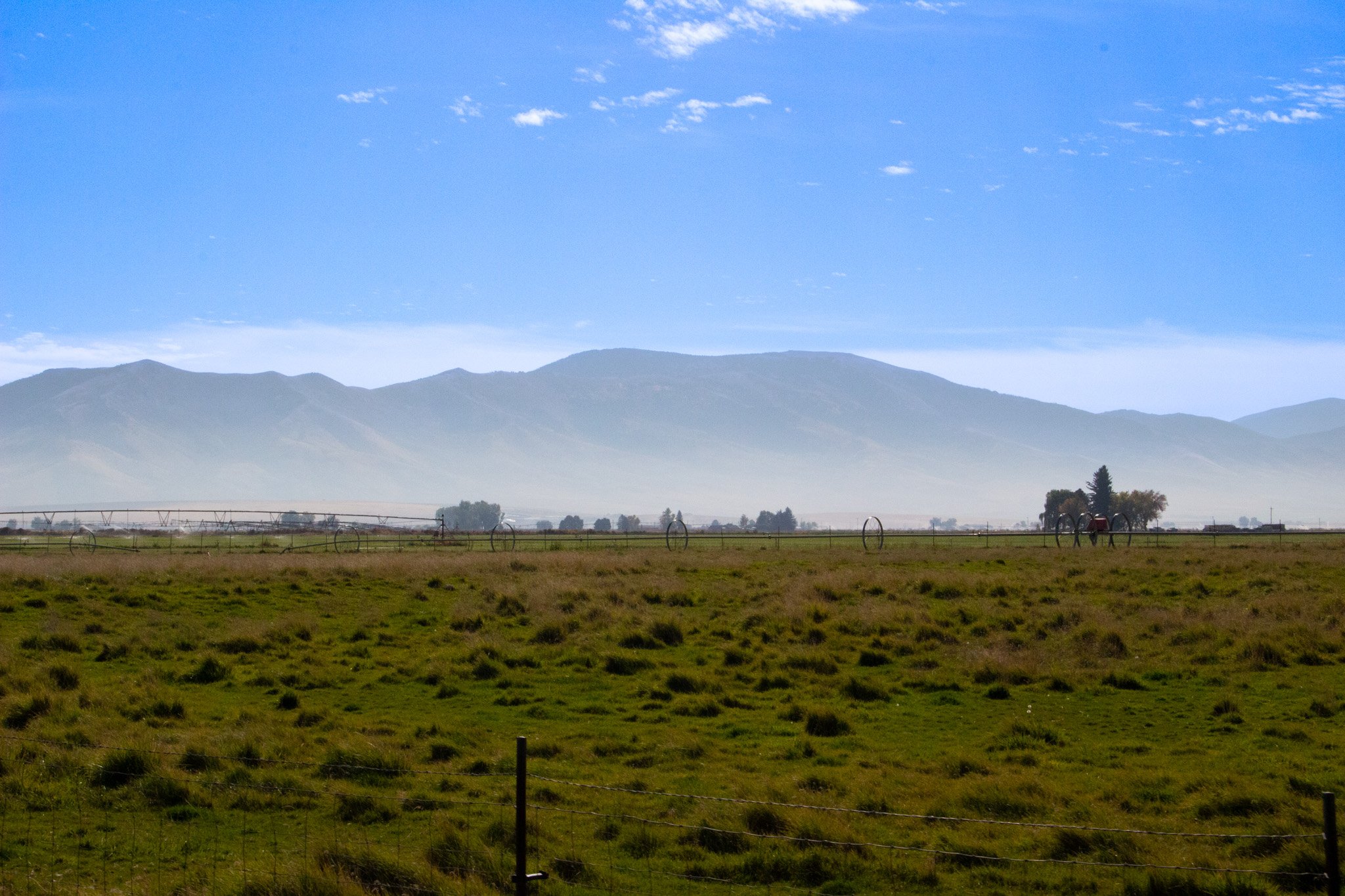 Scenic landscape showing green fields, a distant mountain range, clear blue sky with scattered clouds.