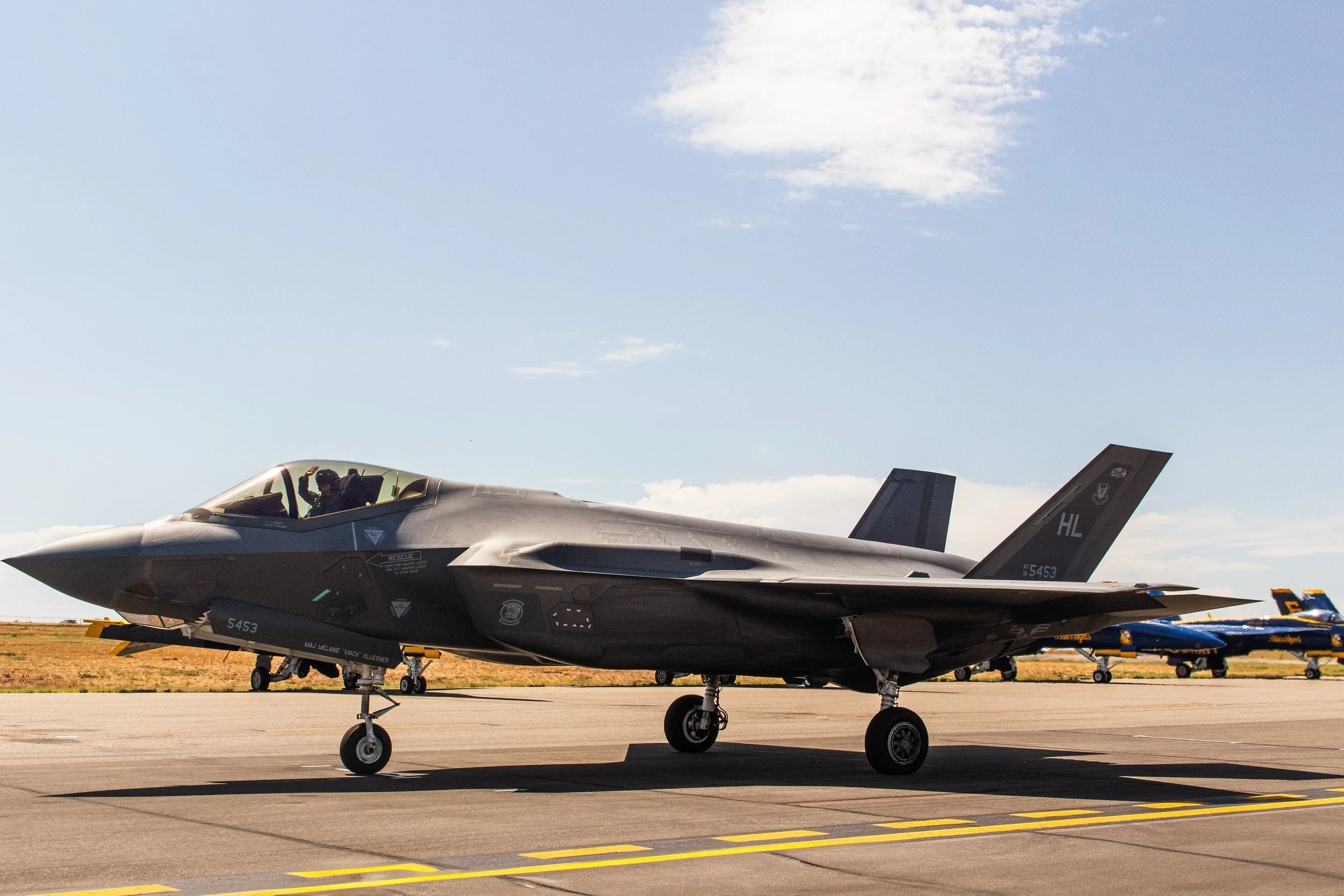 A fighter jet aircraft parked on the tarmac at an airfield with blue sky and clouds overhead. There are additional aircraft visible in the background.