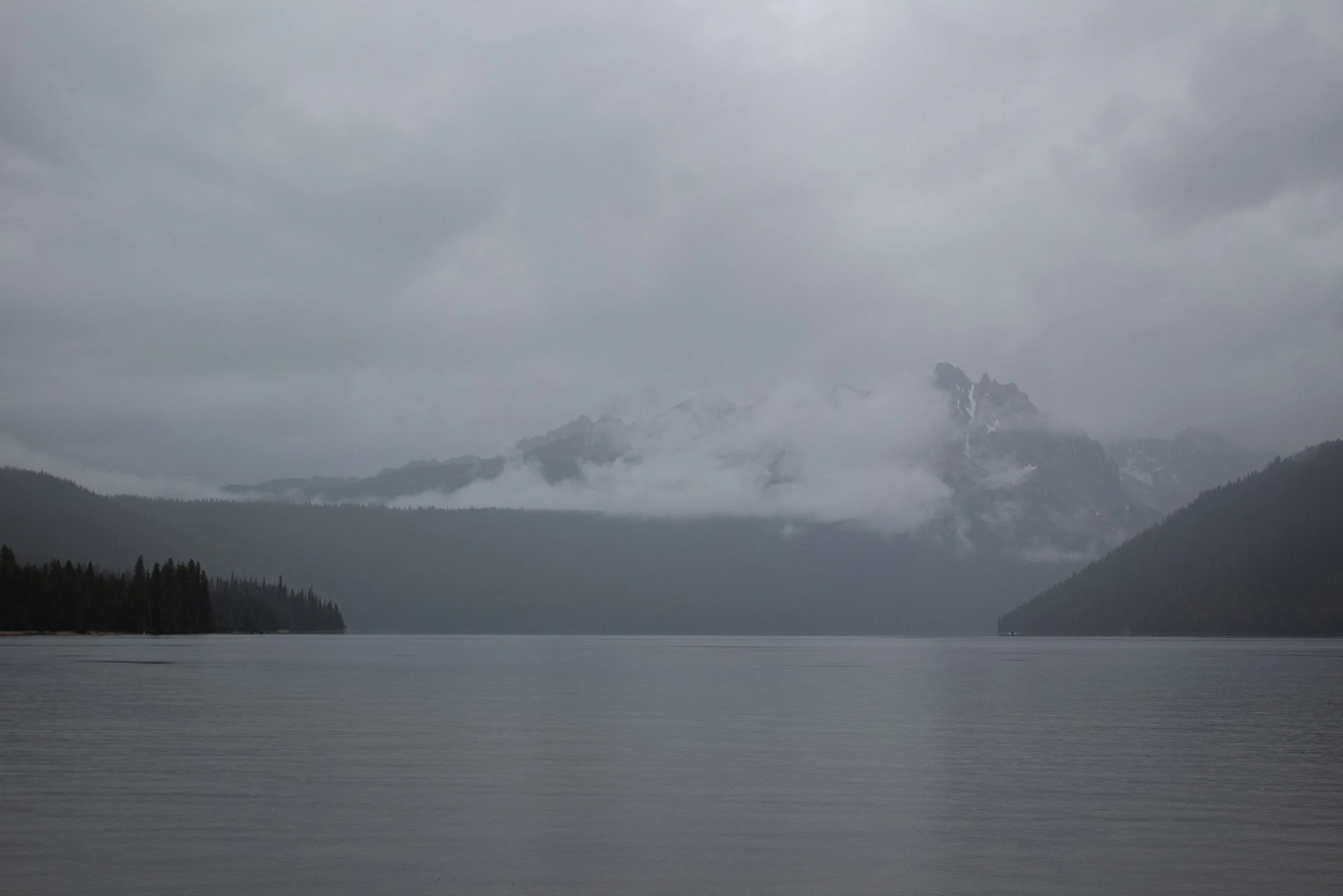 A large body of water with distant mountains partially covered in snow and clouds, surrounded by forested land under a cloudy sky.