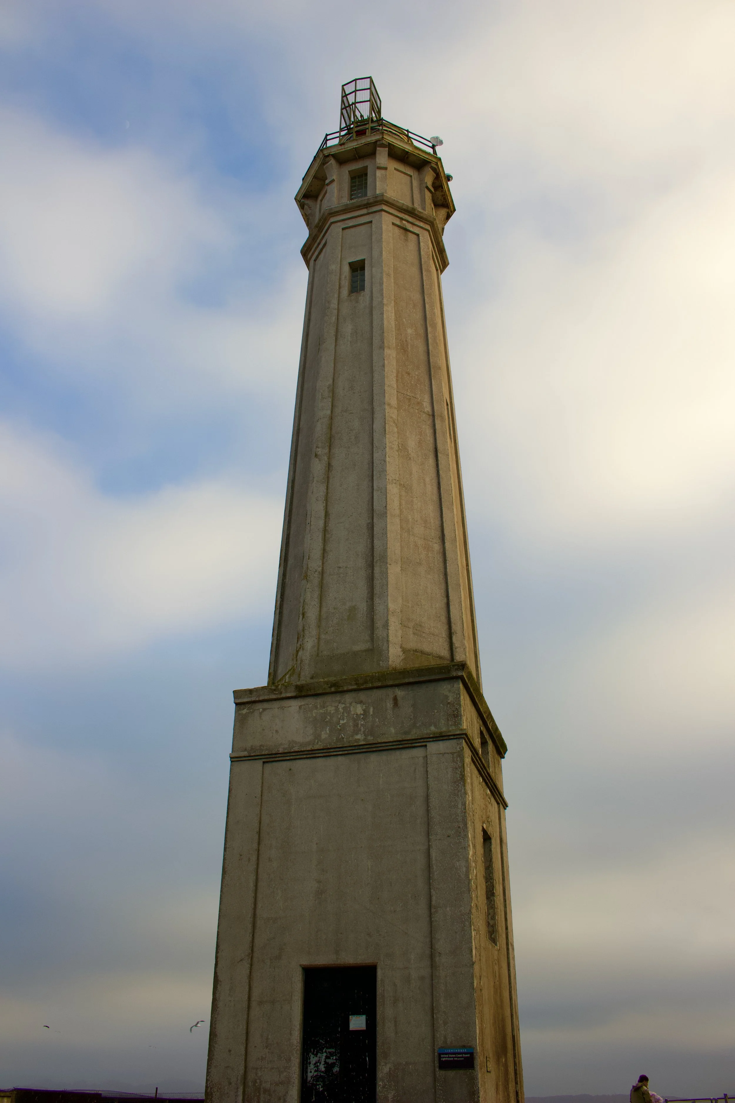 A tall lighthouse with an octagonal top, set against a cloudy sky with a small crescent moon visible. A person stands near the base, looking toward the ground, with seagulls flying nearby.