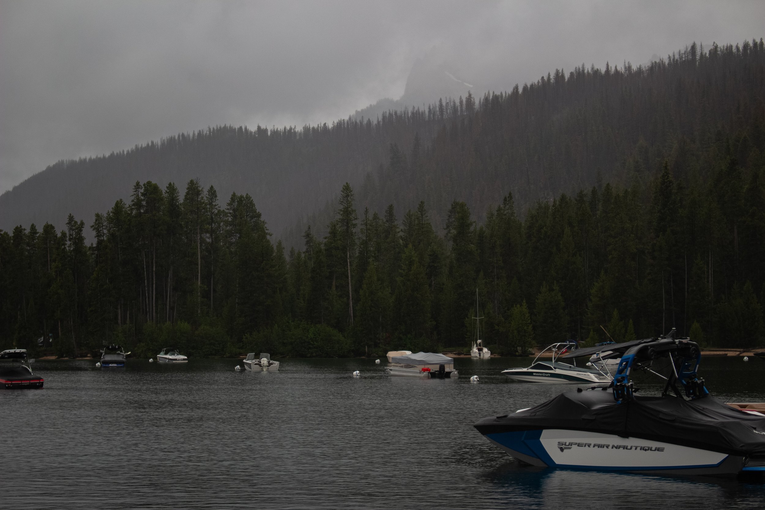 A lake with several boats anchored near the shore, surrounded by forested mountains under a cloudy sky.