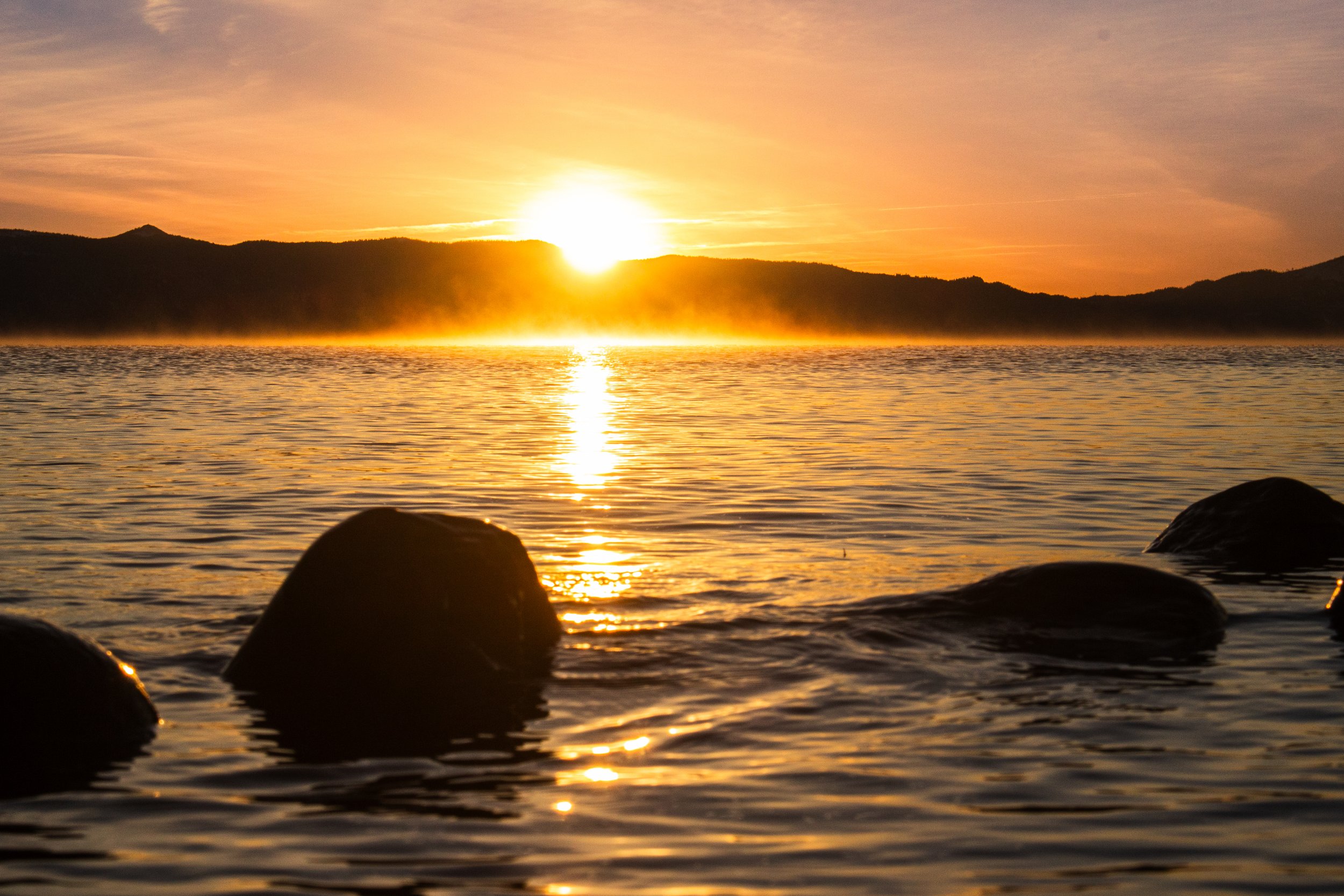 Sunset over a calm body of water with silhouetted rocks in the foreground and a distant mountain range under a sky with light clouds.