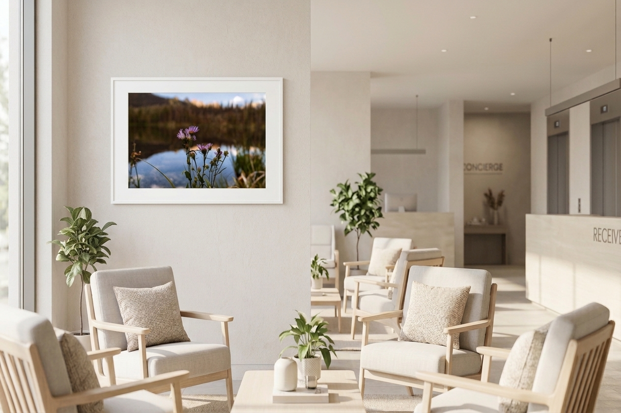 A waiting area with beige armchairs and throw pillows, potted plants, a wall art photograph of flowers, and a reception desk in the background.