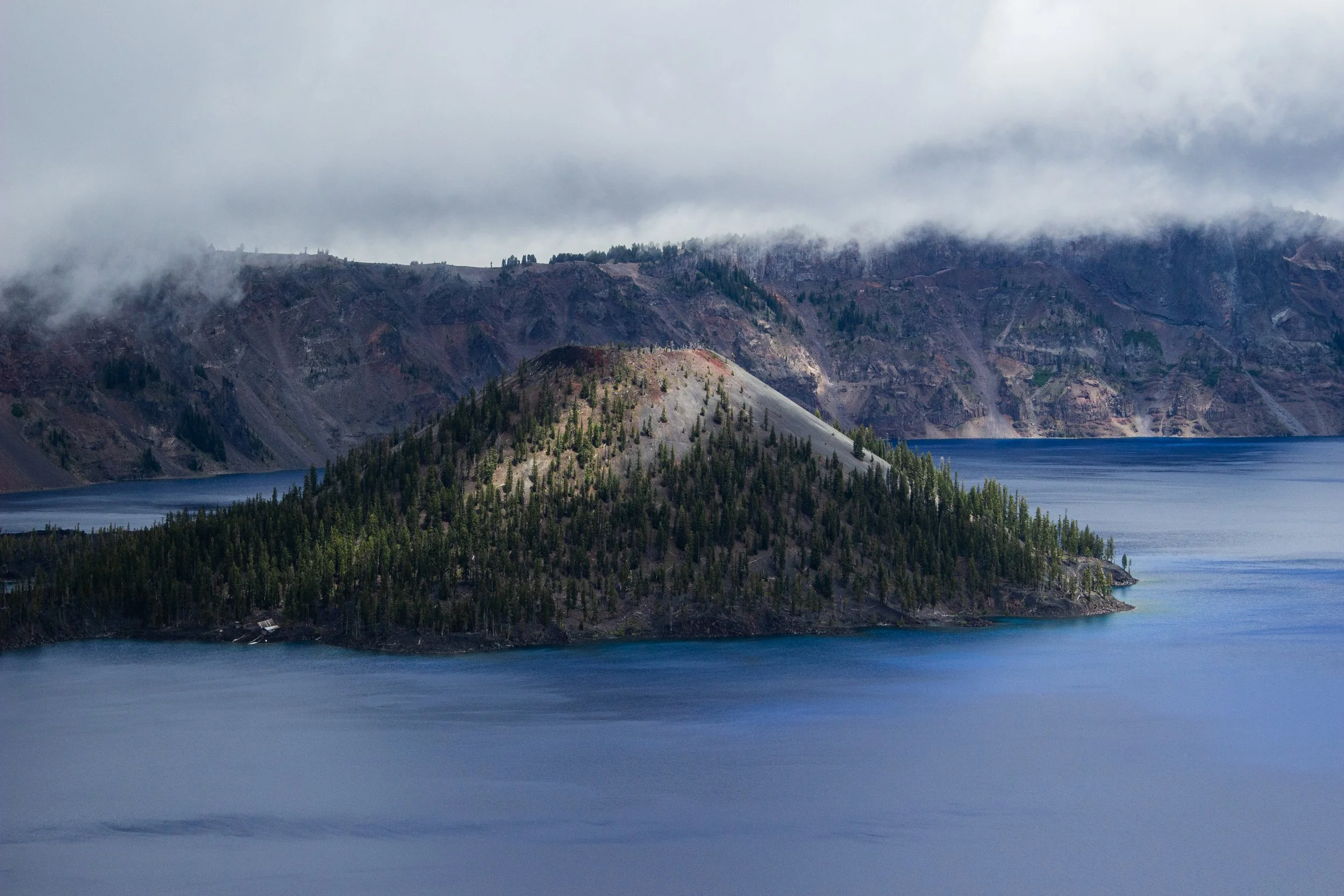 A mountainous landscape with a lake in the foreground, surrounded by evergreen trees. The mountains are rugged with patches of trees and rocky slopes, and are partially covered by low-hanging clouds or fog.