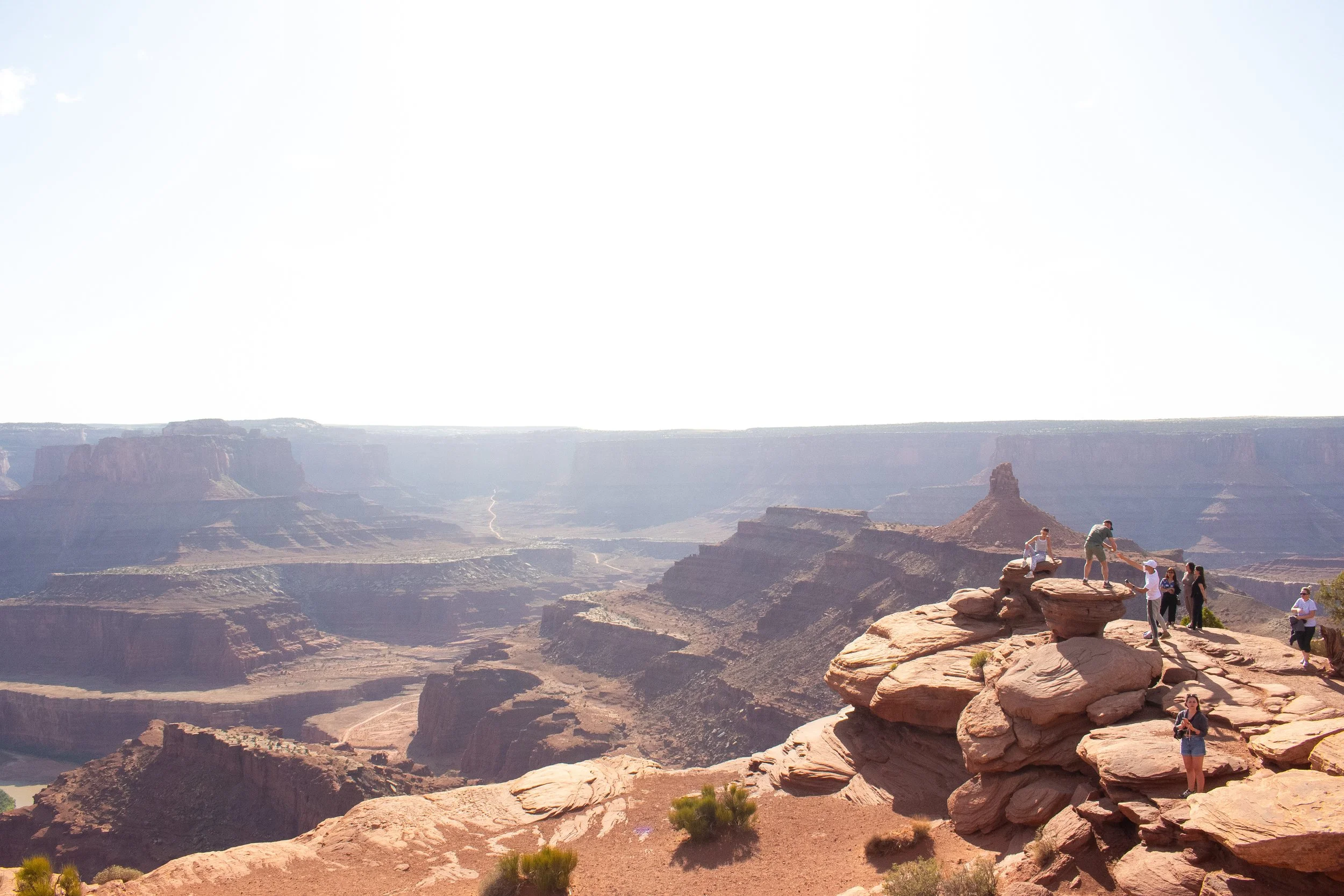 Tourists standing on rocks at the Grand Canyon overlooking the canyon's layers of red and orange rock formations with distant cliffs under a bright sky.
