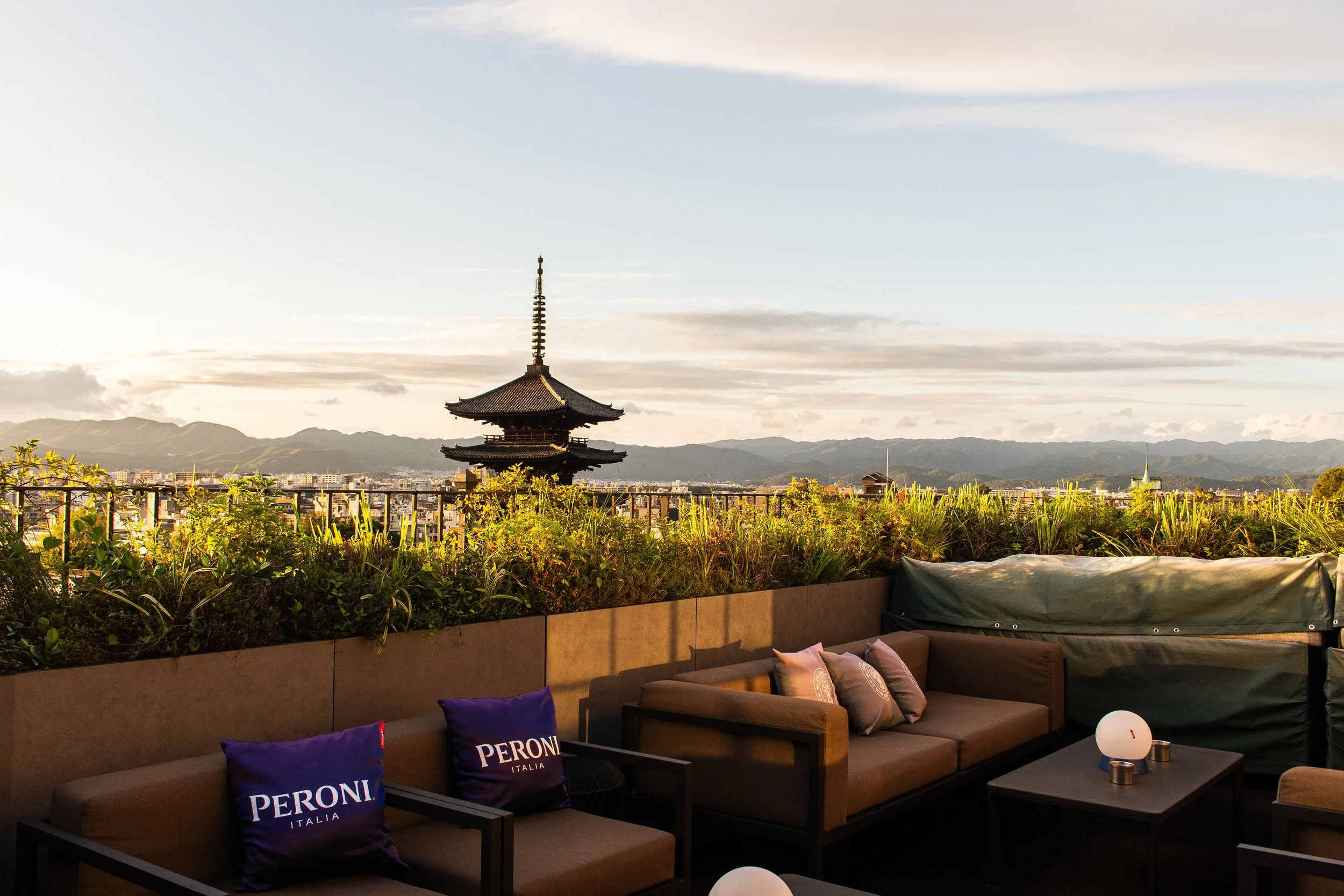 A rooftop terrace overlooking a cityscape with mountains in the distance. The terrace is decorated with a brown sofa adorned with Peroni Italia pillows, small tables with white spherical lamps, and couch cushions in purple, beige, and pink. The background features a traditional pagoda-style building and a clear sky with light clouds.
