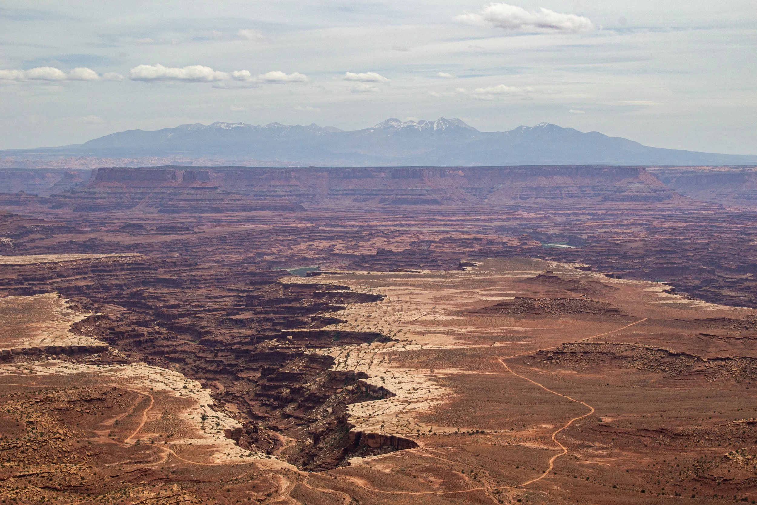 A vast desert landscape with canyons and plateaus, mountains with snow in the distance, and a cloudy sky overhead.