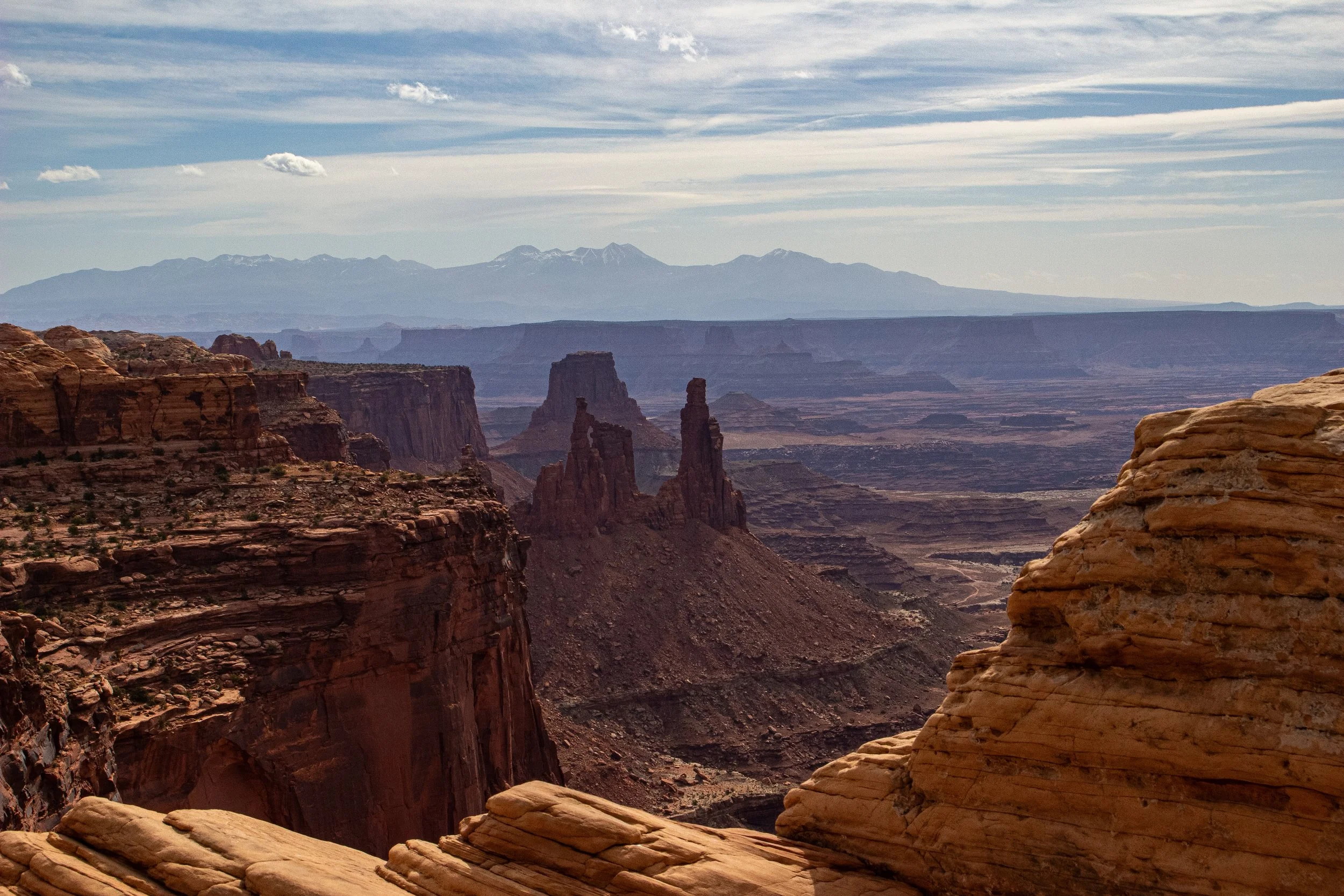 A vast canyon landscape with layered rock formations, cliffs, and spires, under a partly cloudy sky, with snow-capped mountains in the distance.