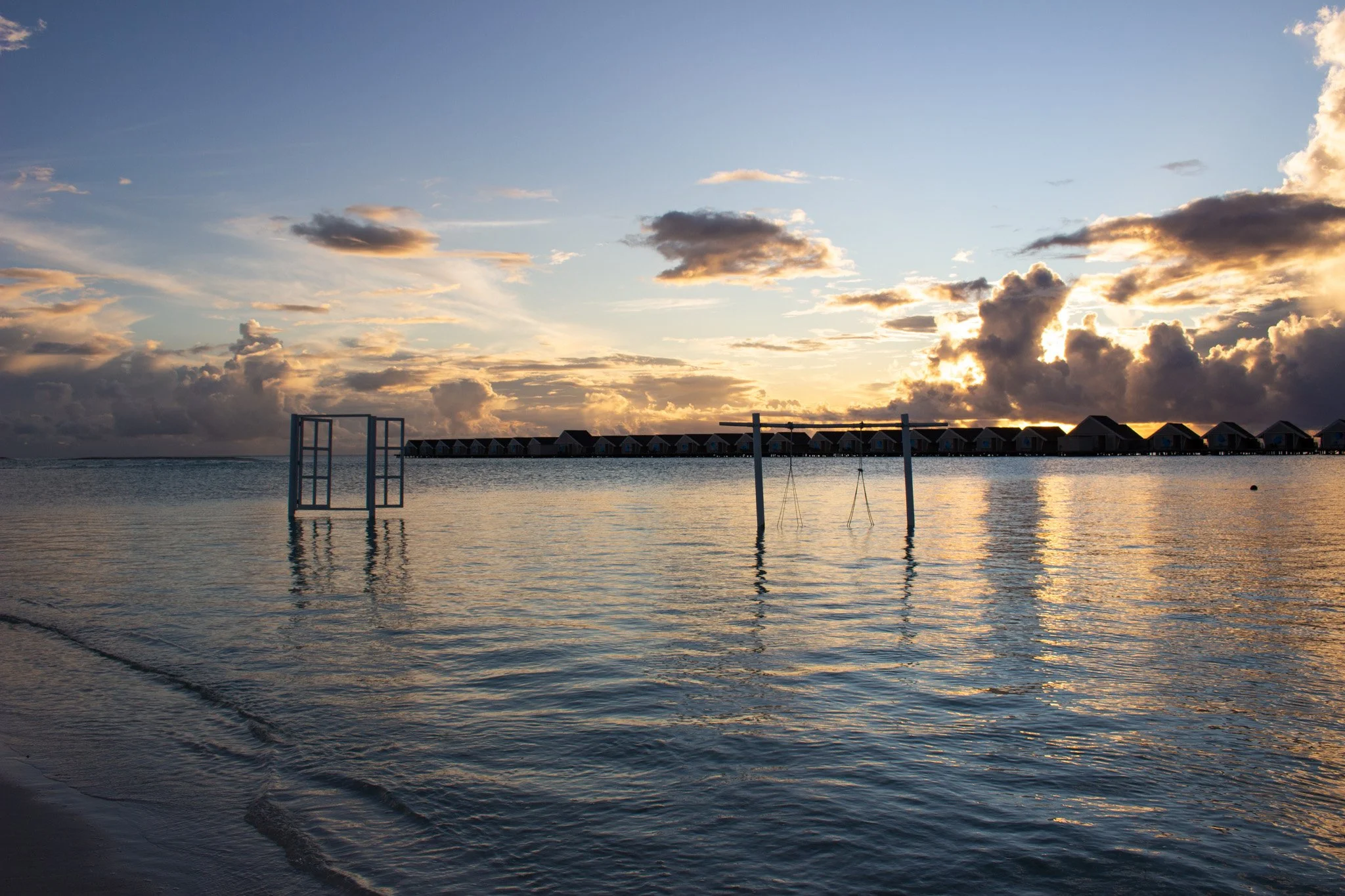 A sunset over water with clouds in the sky, and overwater bungalows in the distance.