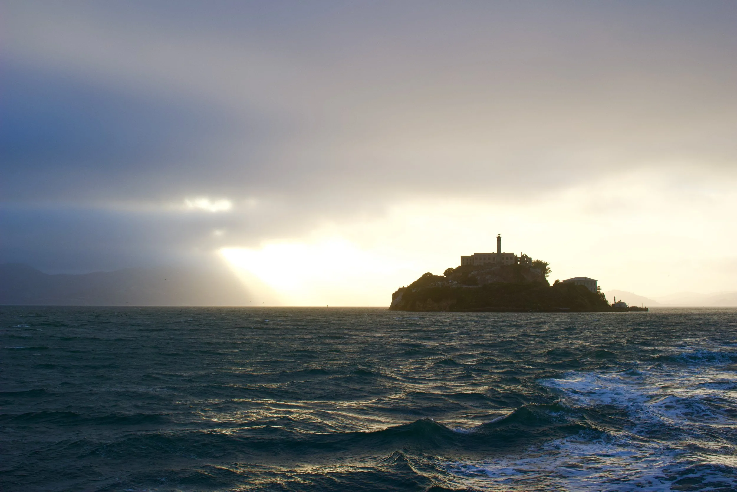 An island with a lighthouse and building structures on top, surrounded by ocean water with gentle waves, under a cloudy sky with sunlight breaking through.