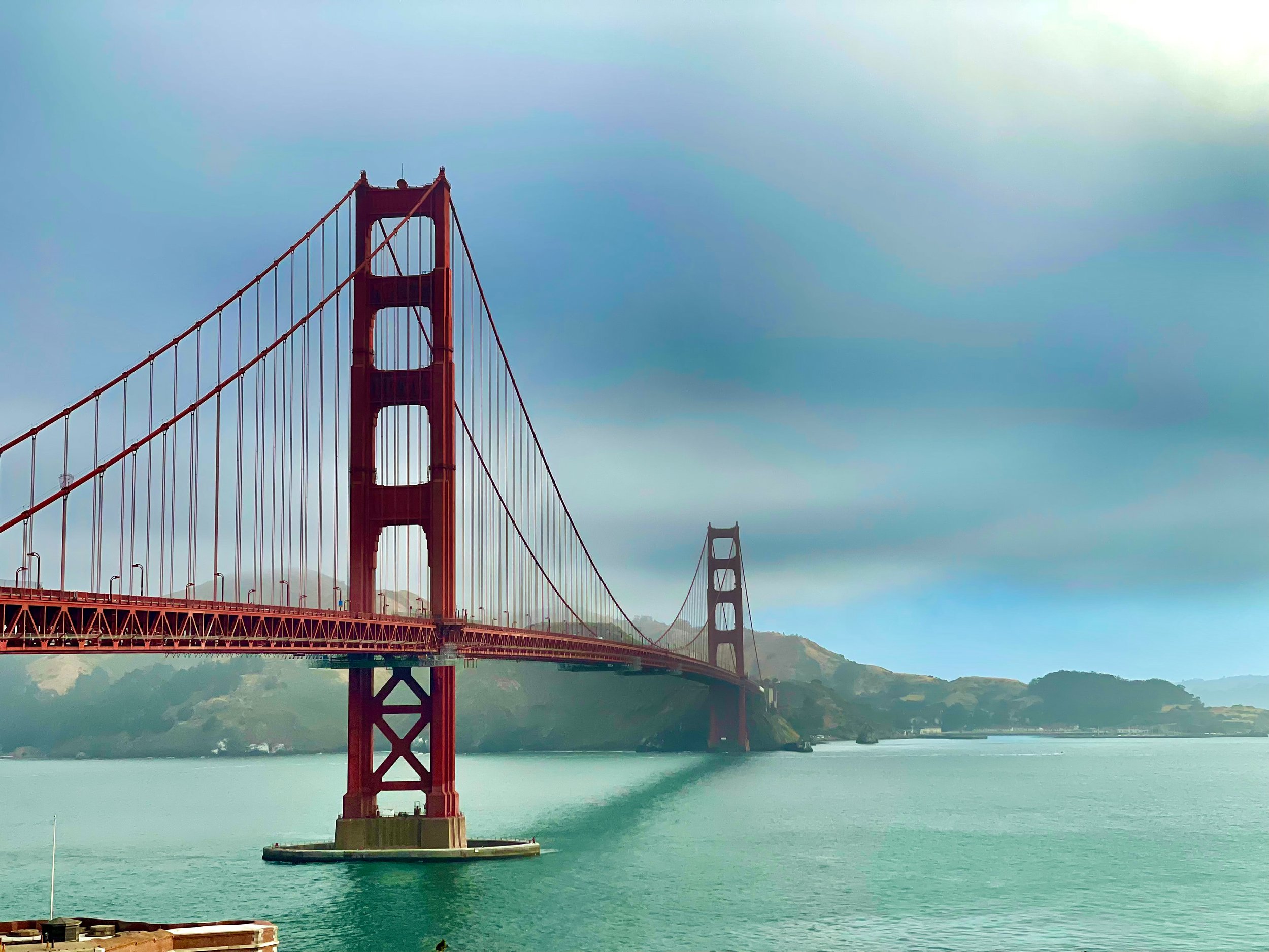 View of the Golden Gate Bridge in San Francisco, California, with the bridge's red-orange towers and suspension cables visible over the water, under a partly cloudy sky.