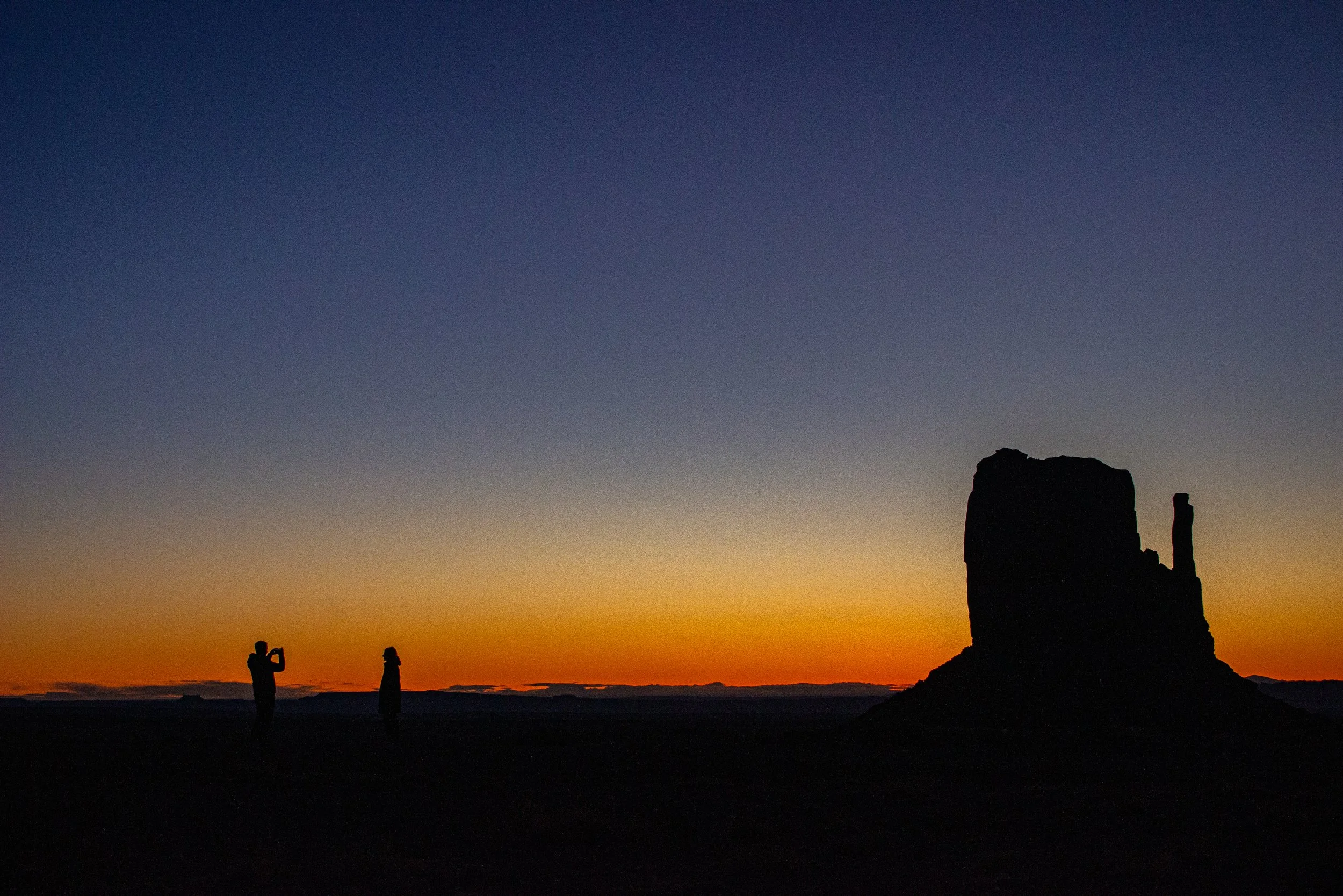 Silhouettes of people taking photographs in front of a sunset with rock formations in the background.