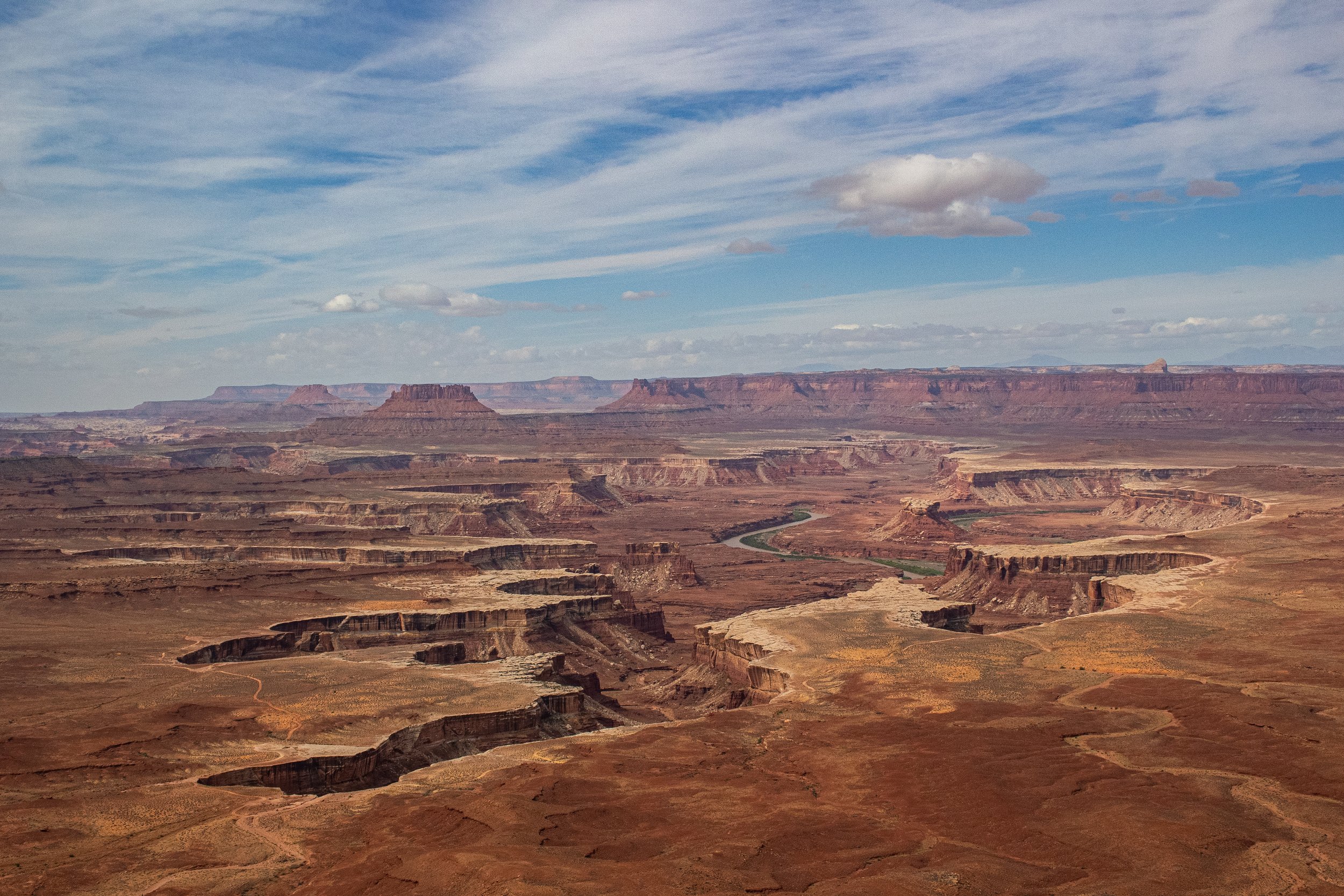 Wide view of the Grand Canyon with layered red and brown rock formations, a winding river at the bottom, and a partly cloudy sky overhead.