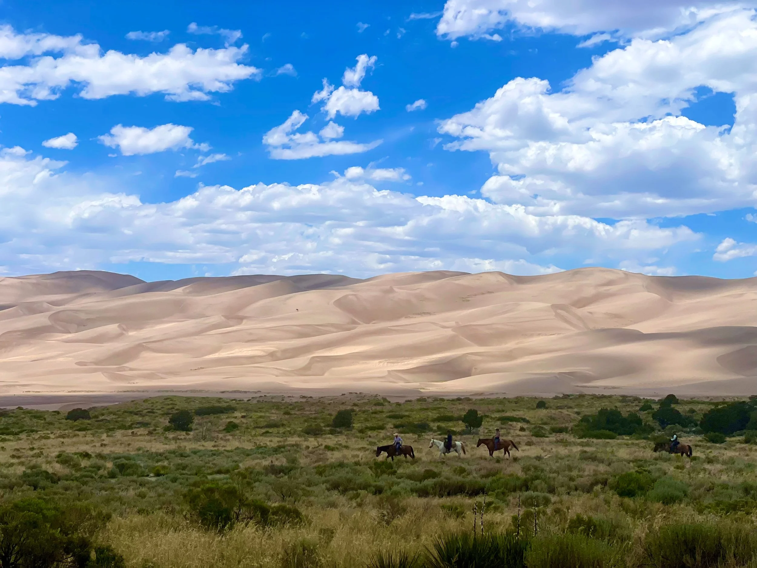 Scenic view of rolling sand dunes under a bright blue sky with scattered white clouds, with a group of horseback riders in the foreground.