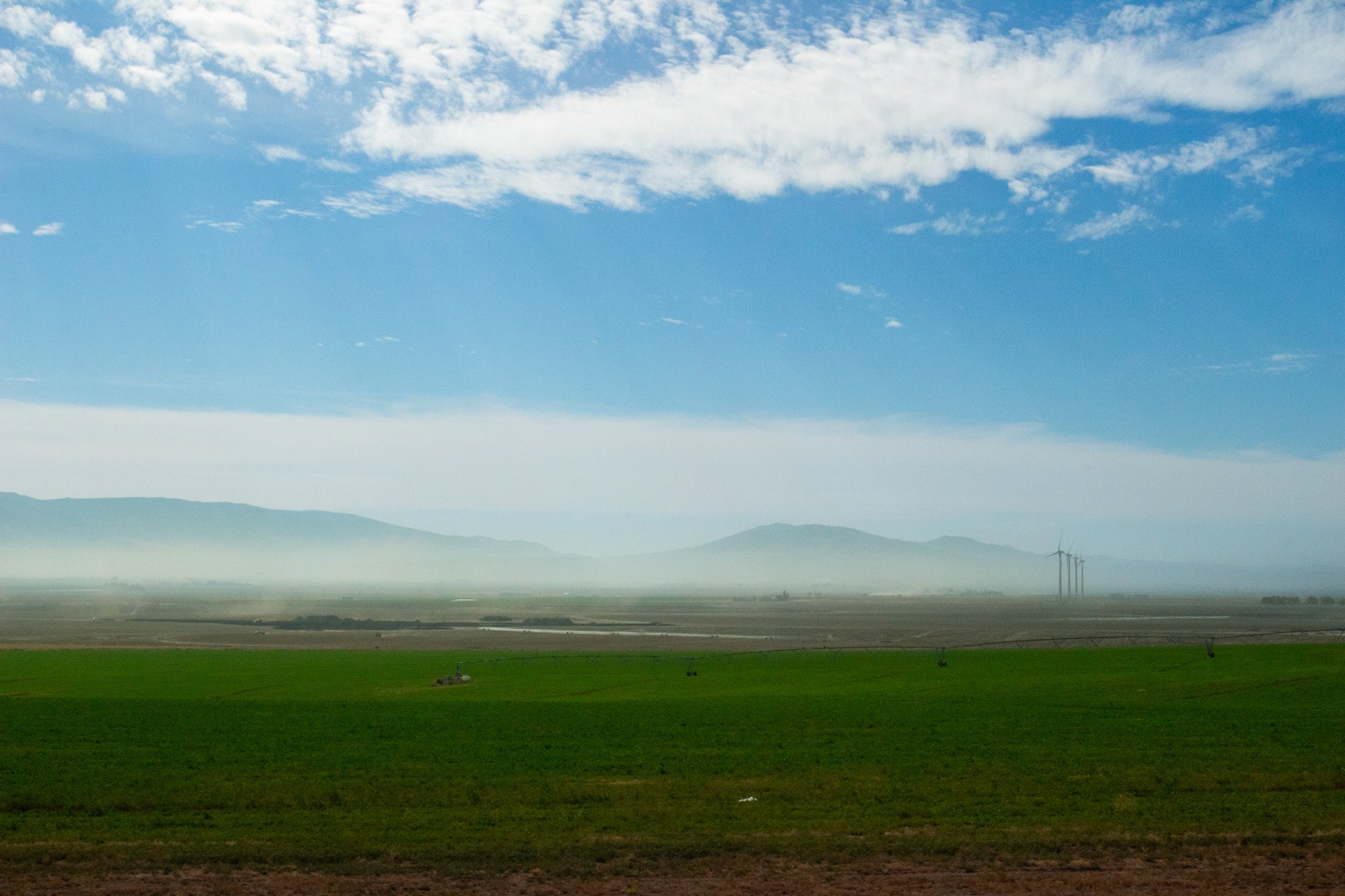 Open landscape with green fields, hazy mountains in the distance, and a blue sky with scattered white clouds.