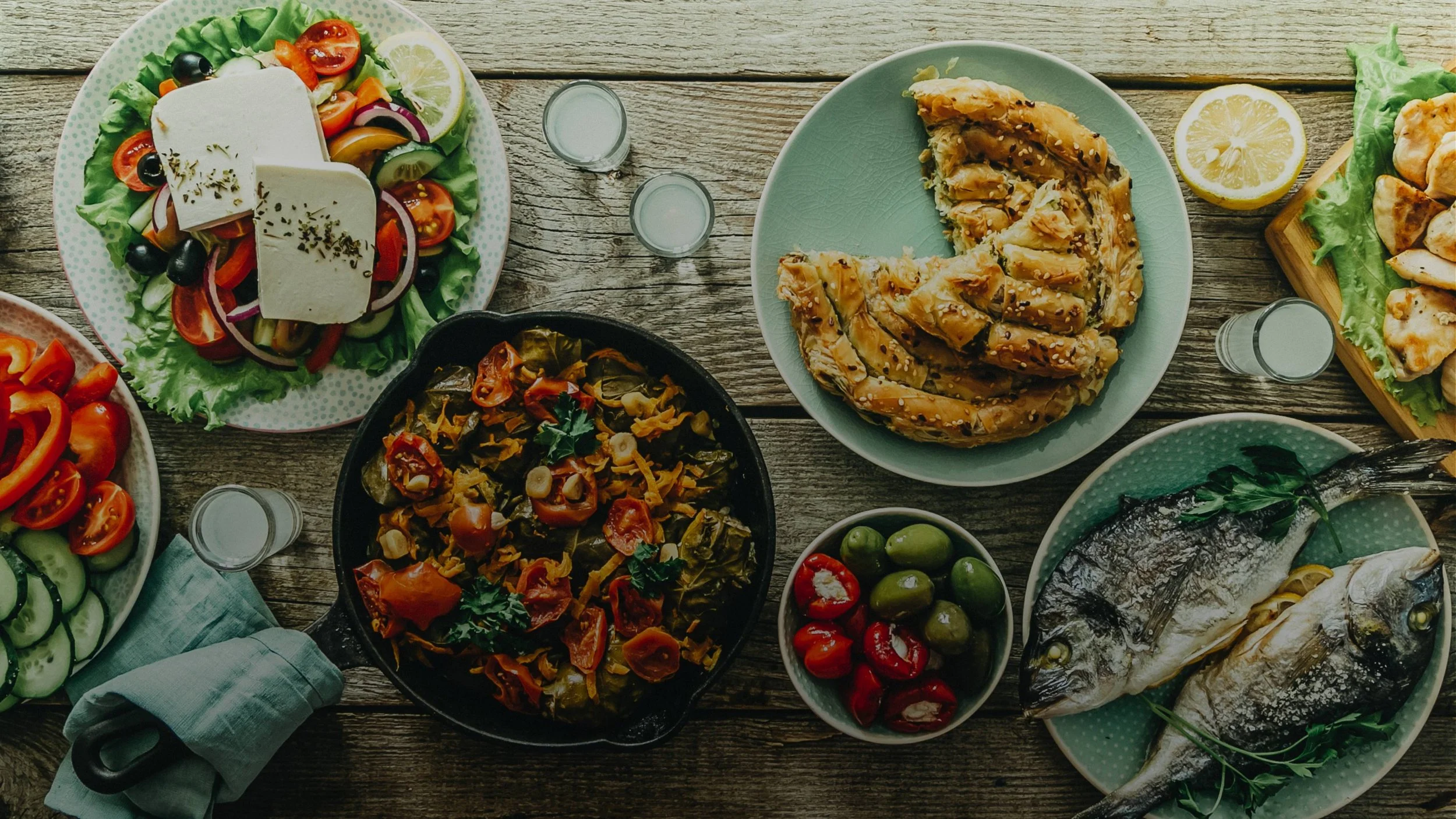 Assorted dishes of grilled fish, salad with cheese, roasted vegetables, olives, and lemon on a rustic wooden table.