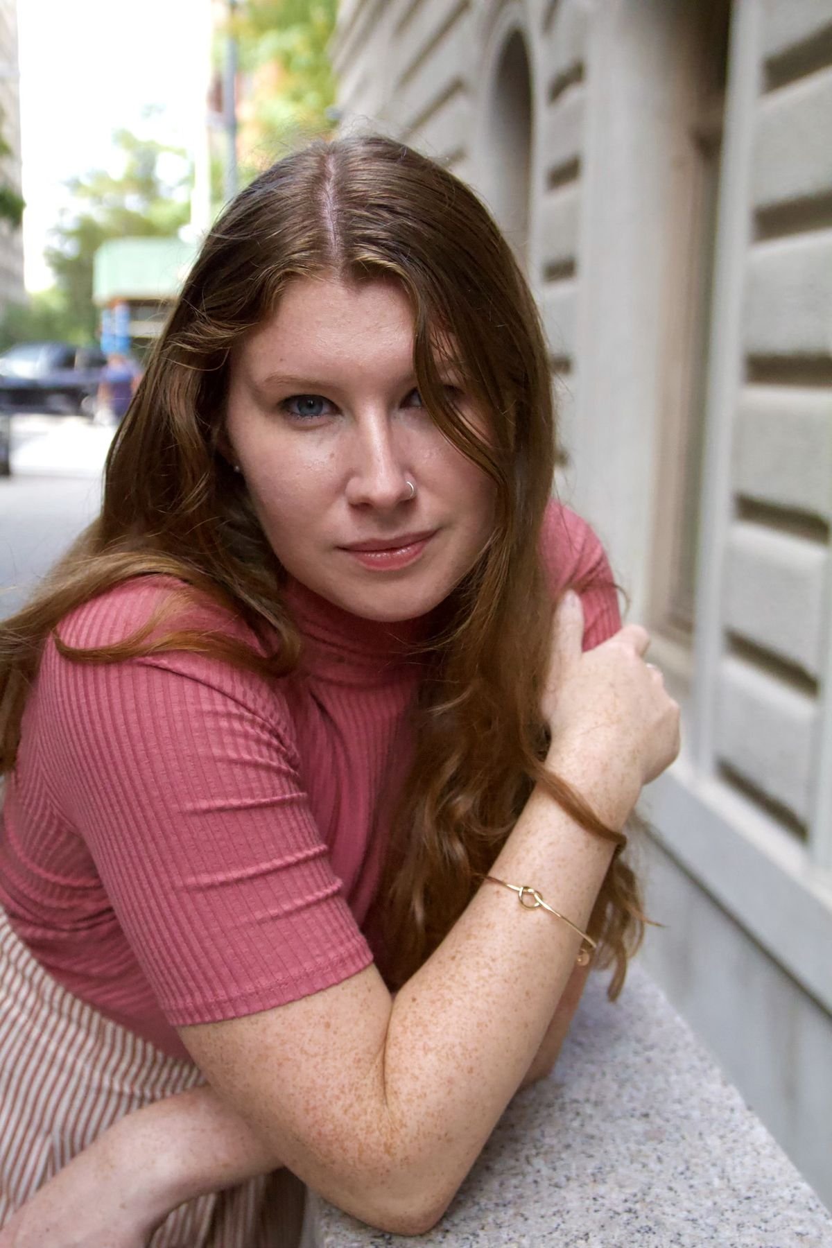 A young woman with long, curly red hair, blue eyes, and freckles, wearing a pink ribbed shirt with short sleeves and a gold bracelet, sitting outdoors at a table with her arm resting on her knees, near a stone building and greenery in the background.