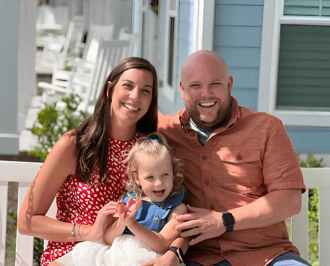 A happy family of three sitting outside on a bench in front of a blue house, smiling and enjoying the sunny day.