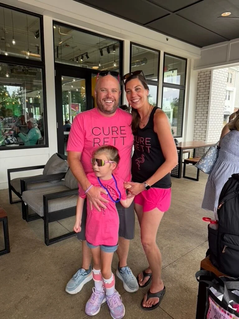 Three people, two adults and a child, standing inside a cafe or restaurant. The adults are smiling, and the child is wearing sunglasses and looking to the side. All three are dressed in pink clothing supporting a cause called Cure Rett.