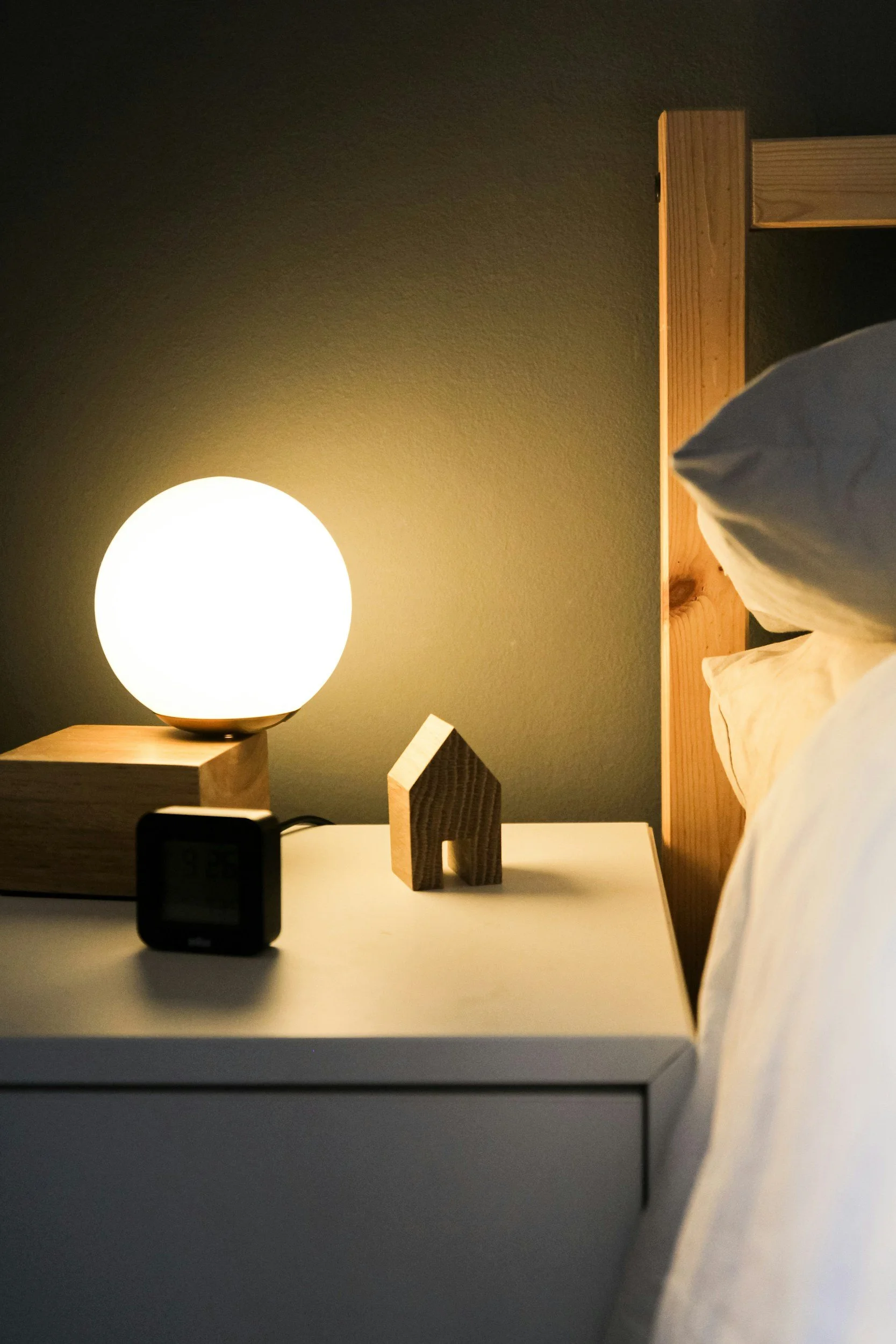 A bedroom scene with a wooden bed frame, a white pillow, and a bedside lamp emitting warm light. On the bedside table, there are a small, black digital clock, a wooden decorative house, and a dark-colored object. The wall behind the bed is plain and dark-colored.