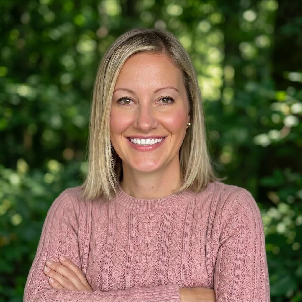A professional woman with blonde hair smiling and crossing her arms, outdoors with green trees in the background.