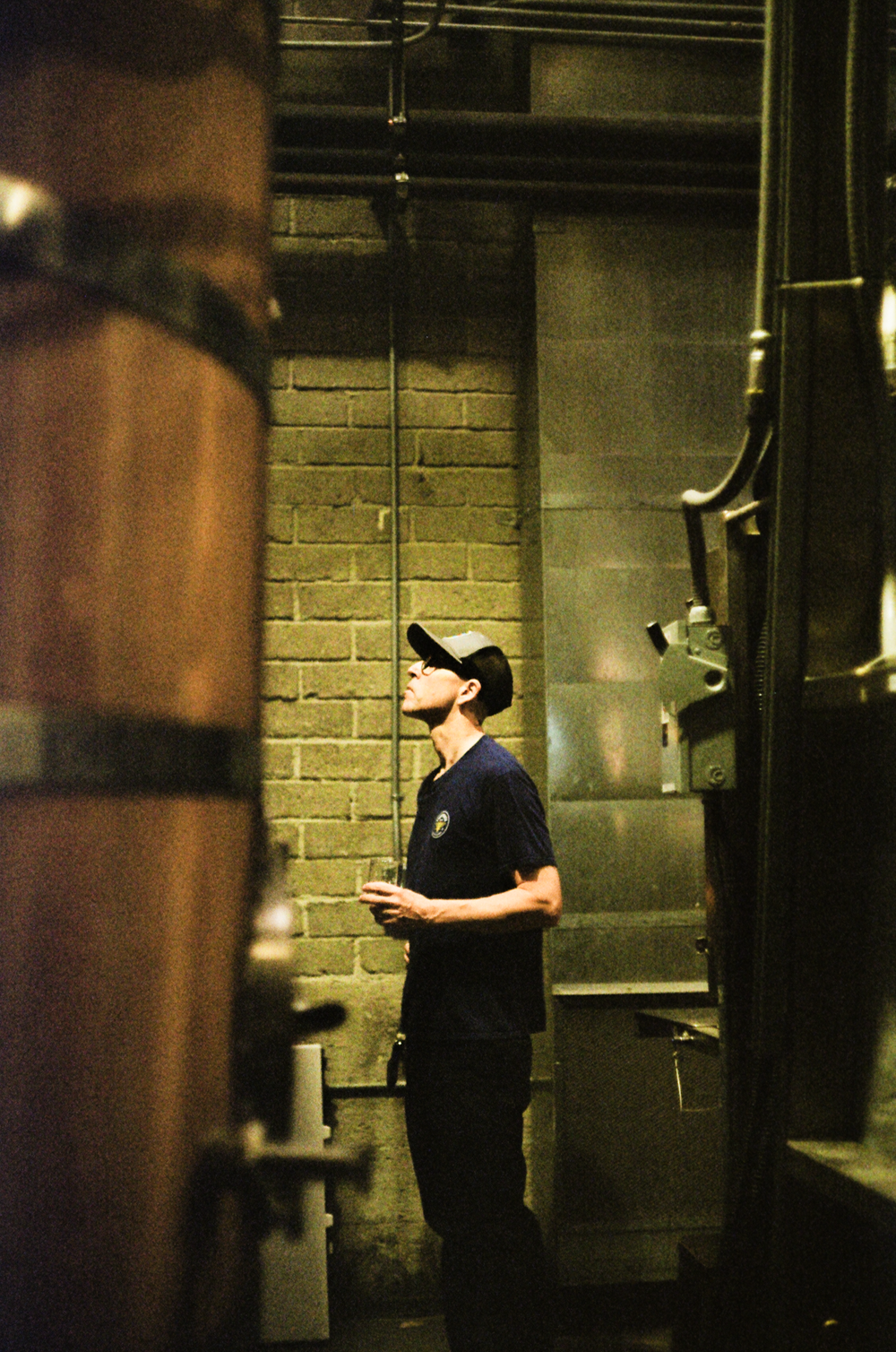 A distiller looking up at the stills between tall barrels at Storyhouse Spirits in East Village, shot on 35mm film