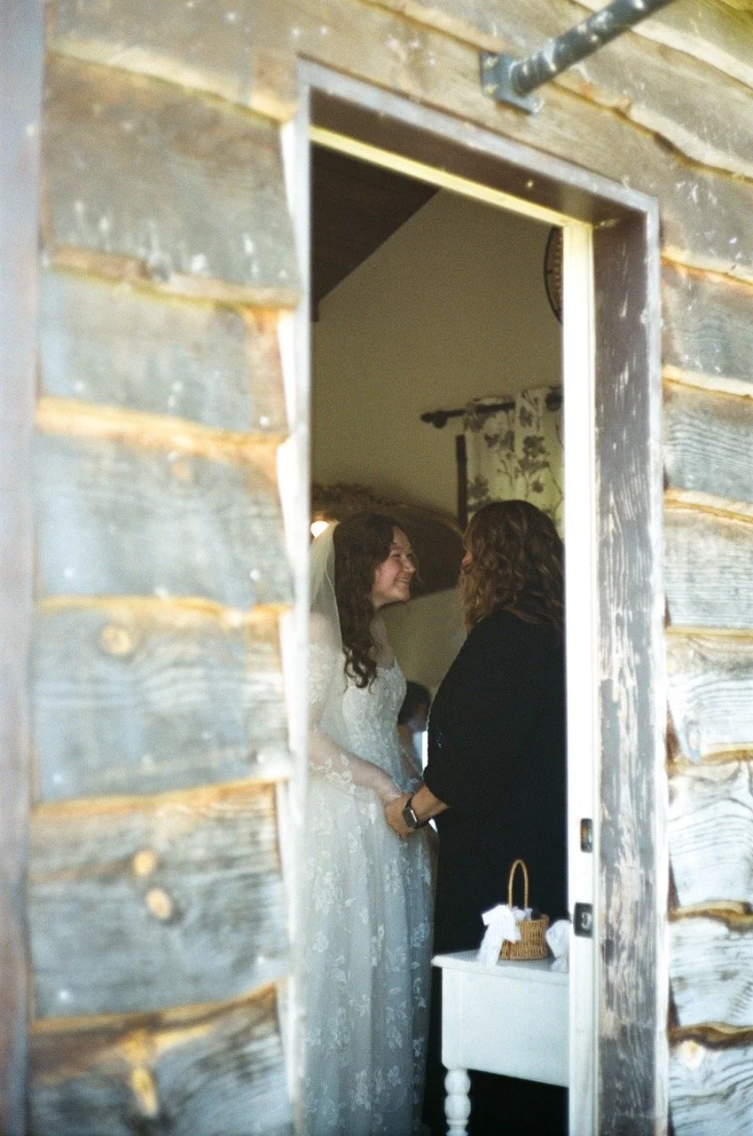 Carley in her wedding dress holding hands with a guest inside a wooden cabin doorway, shot on film