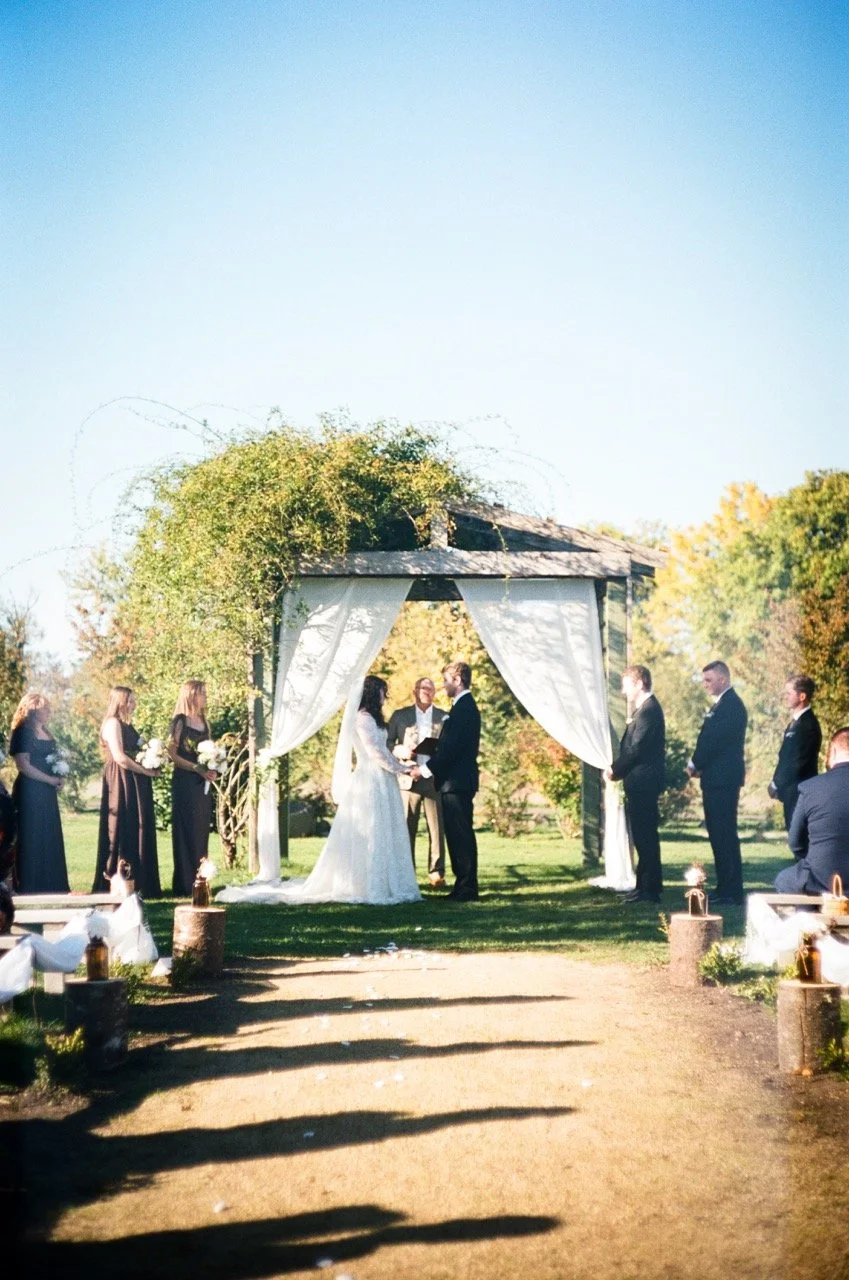 Guests standing along the aisle as the ceremony starts under a white arch at Creekside Farms, shot on film